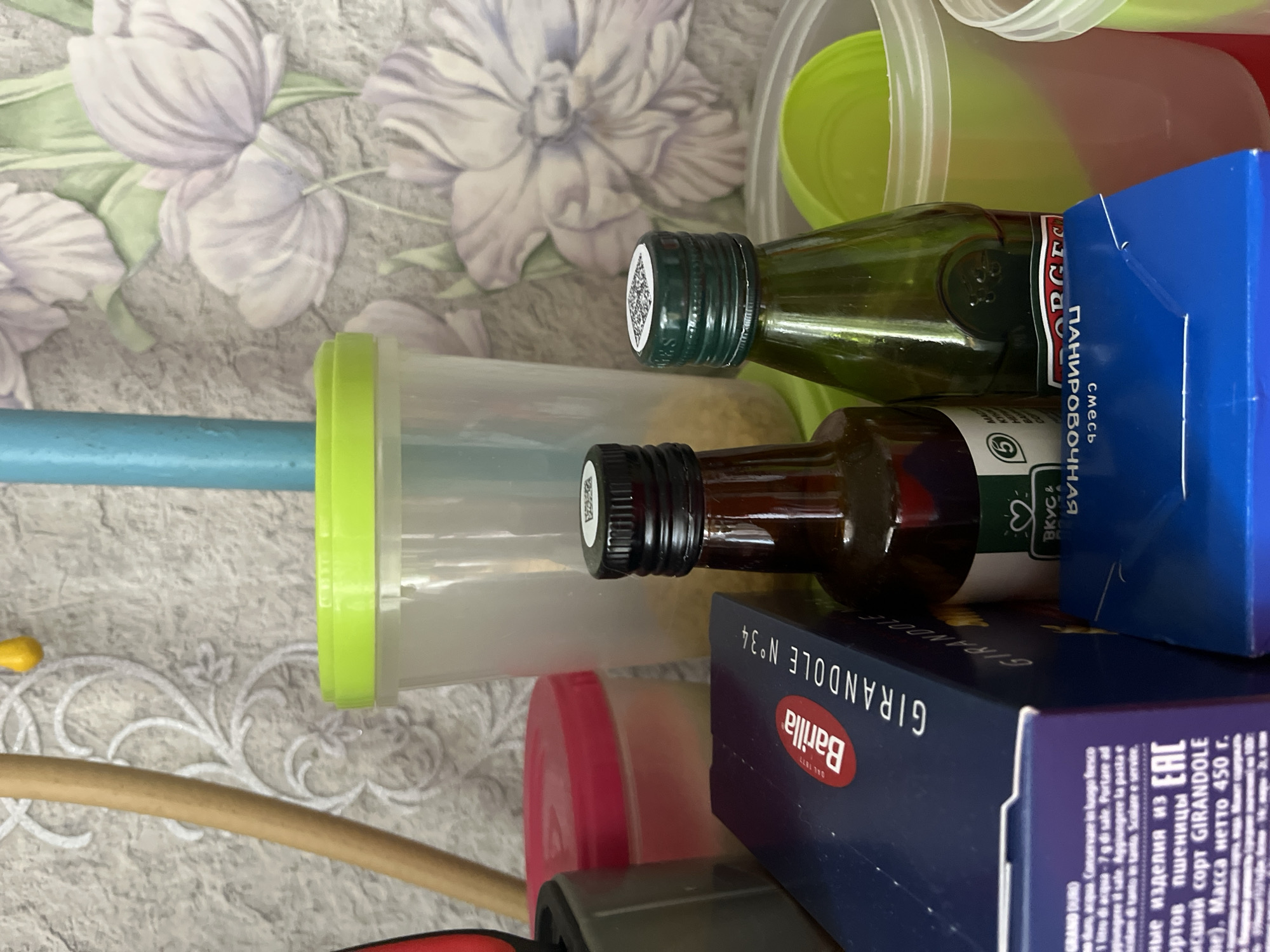 The image shows a still life arrangement of kitchen items. There are two bottles, one with a green cap and one with a black cap. Next to them is a container with a green lid and a blue handle. Additionally, there is a Barilla pasta box (GIRANDOLE N°34) in blue and white, with Russian text, and other colored containers partially visible. The background appears to be a table with a floral patterned covering. The text on the pasta box identifies the product as "GRAND DURO" pasta, and indicates the country of origin is somewhere in the Russian speaking region ("ные изделия из" translates as "articles from") but the text also includes instructions written in Italian. The location is presumed to be Perm, Russia, based on the presence of Cyrillic text.