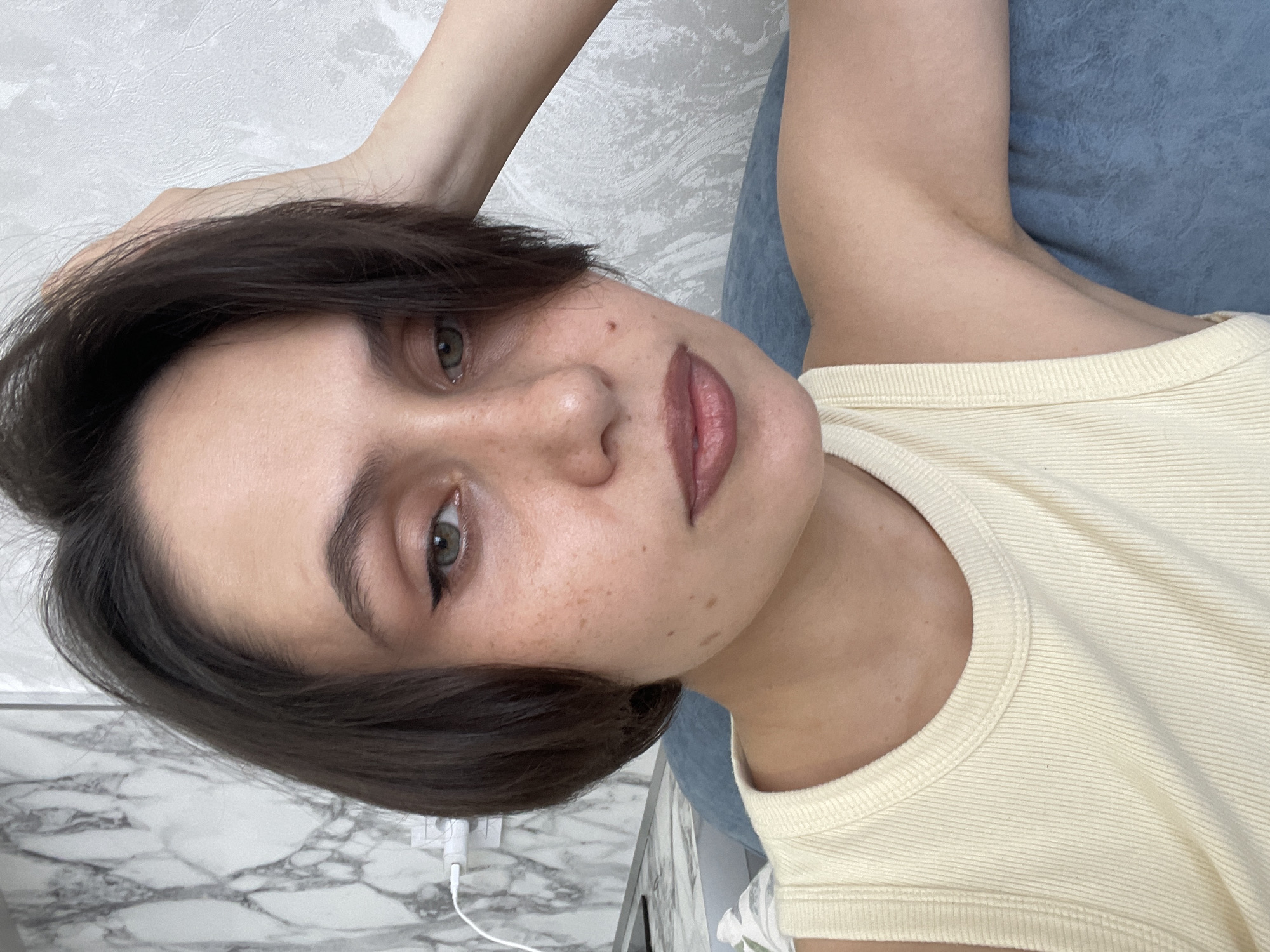 A woman is positioned diagonally in the frame, her head tilted towards the left side. She has fair skin with some freckles, short dark brown hair, and light-colored eyes with eyeliner. She is wearing a light yellow ribbed tank top. Her left arm is raised above her head, and her right shoulder is visible. The background consists of a white textured wall and a blue surface. In the lower left corner, there is a white marble-patterned surface with a white charger and cable. The location is not discernible from the given image, and there is no evidence to suggest it is in Izhevsk, Russia.