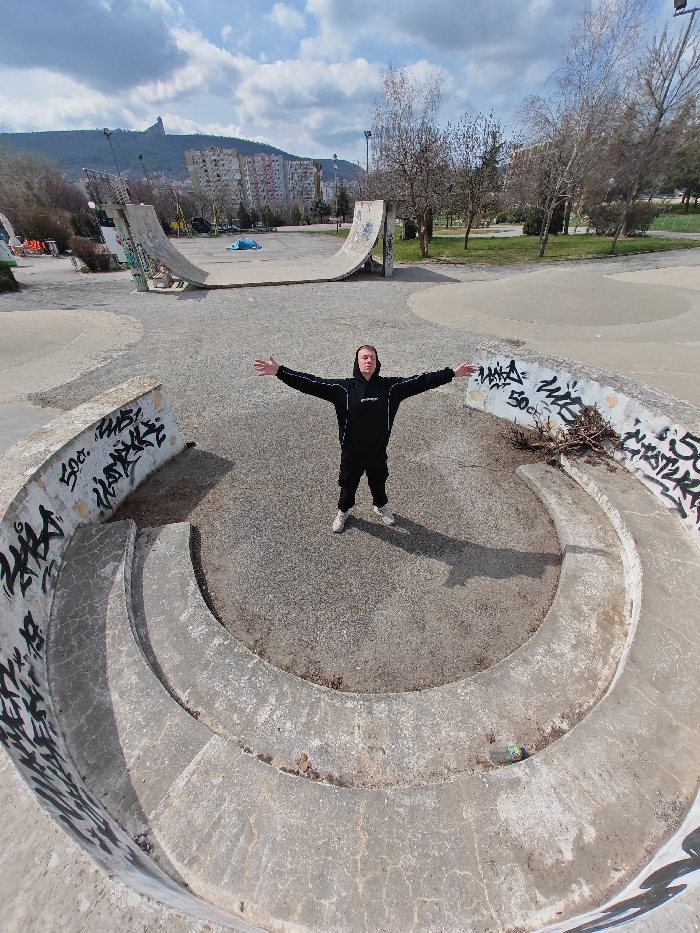 The image displays the Shumen Skatepark in Shumen, Bulgaria, an outdoor concrete facility under a partially cloudy sky. Centrally positioned within a circular, multi-tiered concrete bowl structure, a Caucasian male, approximately 25-35 years old, stands facing the viewer with arms extended horizontally. He is dressed in a black hooded sweatshirt with white stripes on the sleeves, black pants, and white sneakers. The concrete walls of the circular structure are heavily covered in black graffiti. To the man's immediate right, a green plastic bottle lies on the ground within the structure. Further into the skatepark, two prominent concrete ramps or half-pipes are visible. The background features multiple multi-story residential buildings, characteristic of urban architecture, with a rugged mountain peak, part of the Shumen Plateau, rising distinctly in the far distance on the left. Sparse, bare deciduous trees and patches of dry grass are distributed throughout the park. The daylight conditions cast a discernible shadow of the man to his left.