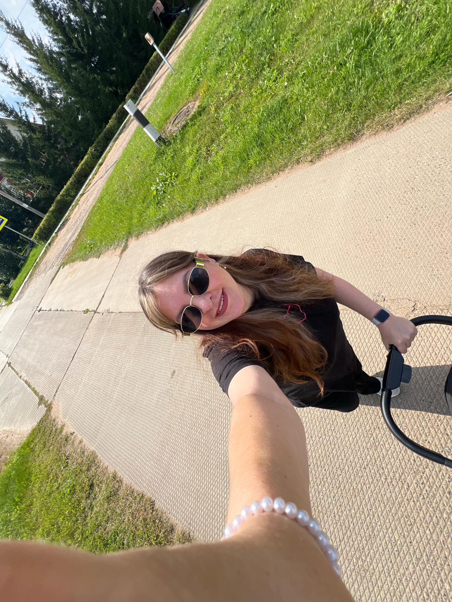 A woman with long brown hair and sunglasses is taking a selfie while pushing a stroller. She is wearing a black t-shirt and a pearl bracelet on her left wrist. The stroller has a black frame and a black handle. They are on a paved path in what appears to be a park or a residential area. To the left of the path is a grassy area with trees and a hedge. In the background, there is a road and some buildings. The scene is illuminated by bright sunlight, casting shadows on the ground. The location is identified as Noviy, Russia.