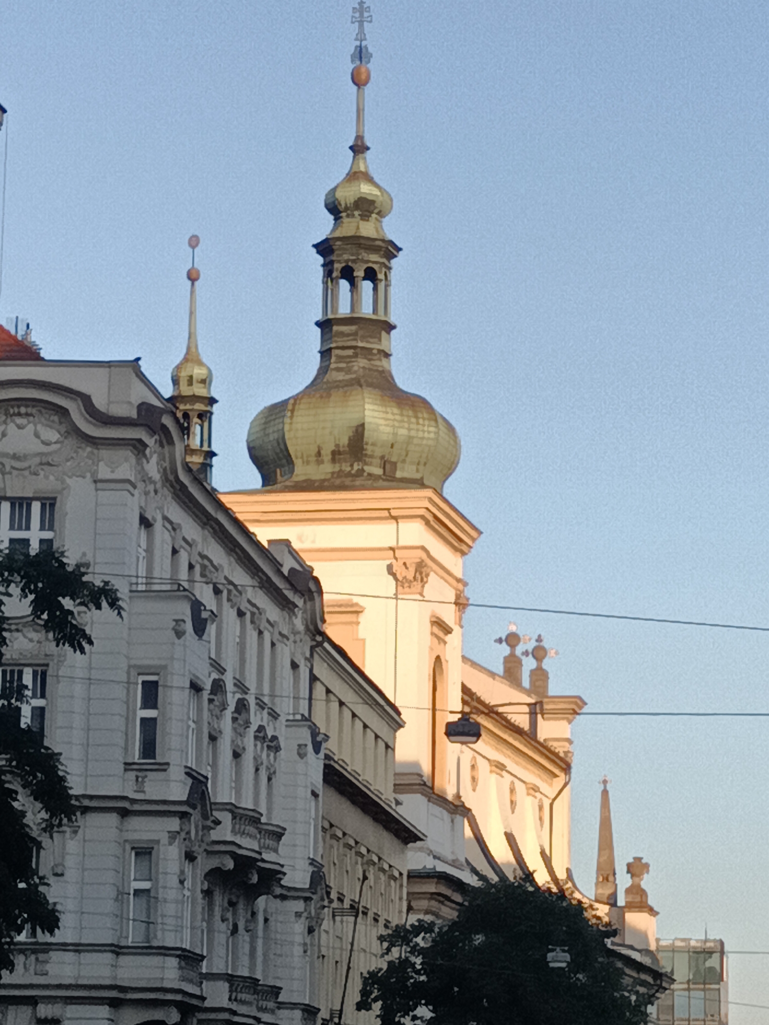 The image depicts an urban scene in Prague, Czechia, focusing on the architecture of buildings and church towers against a clear sky. In the foreground, the left side of the frame shows a grey, ornate building with multiple windows and balconies. To its right, a prominent, multi-tiered church tower with a green, bulbous dome and a tall spire topped with a cross dominates the central and right portions of the image. Smaller spires and architectural elements are visible on other structures further in the background. The light suggests it is either early morning or late afternoon, casting a warm glow on some of the buildings. The scene is devoid of people and vehicles, emphasizing the architectural elements and the cityscape.