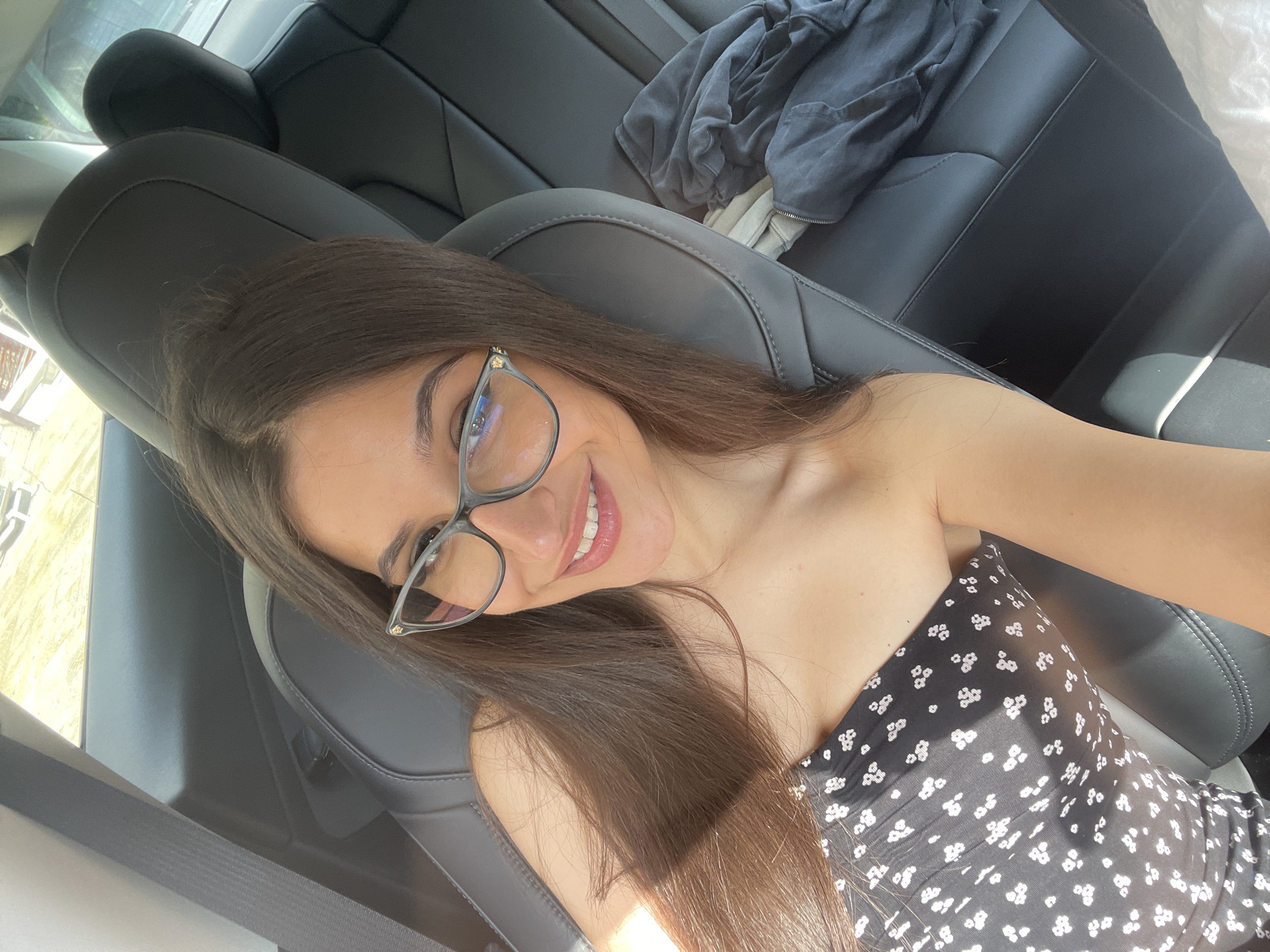 A young woman with long brown hair and glasses is taking a selfie in the back seat of a car. She is wearing a black strapless dress with white floral patterns. The interior of the car is black leather. There are some clothes piled on the seat behind her. The windows are open, and sunlight is streaming in. The background appears to be a suburban or rural area with some trees visible.