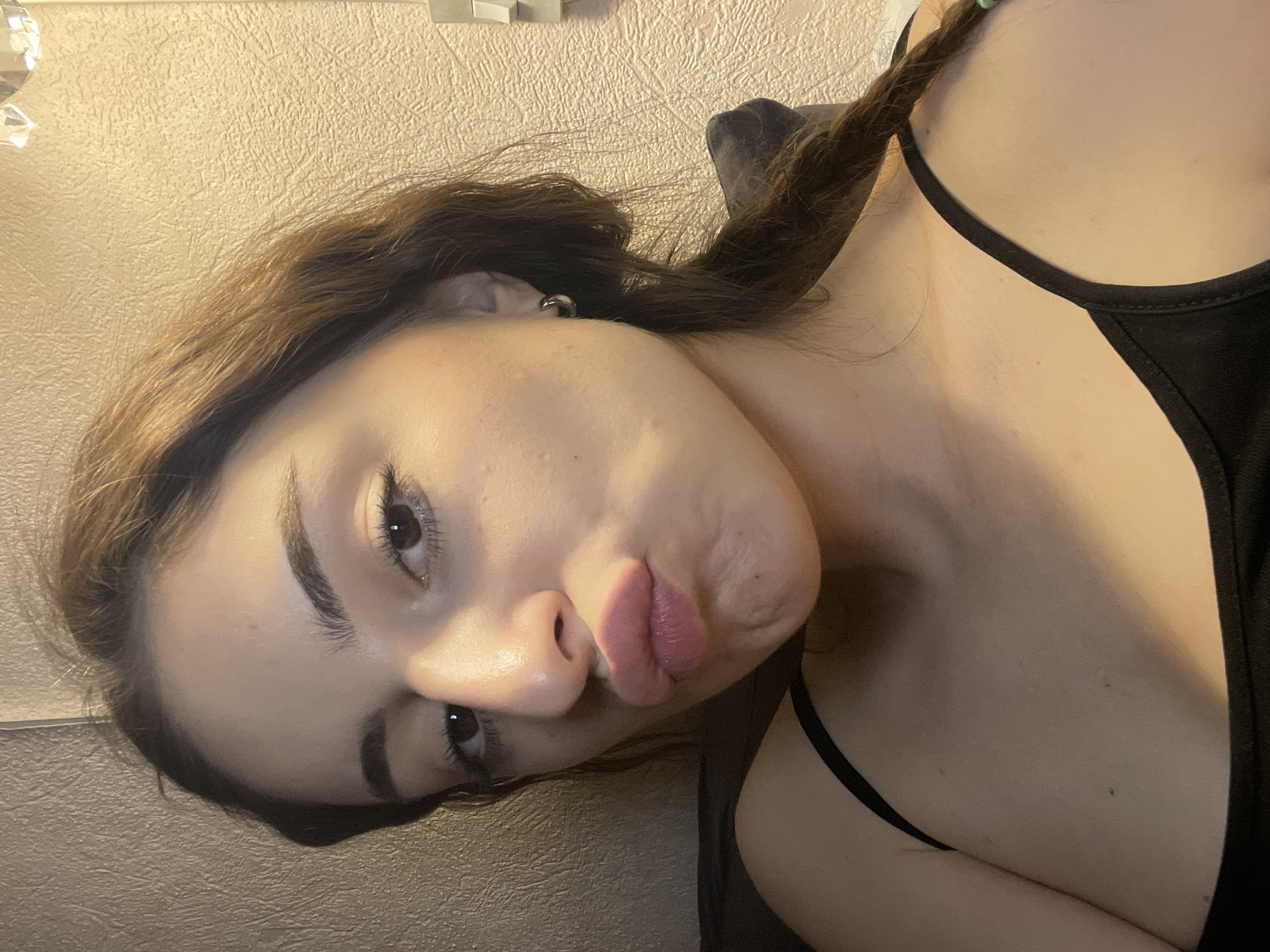 A young woman with brown hair and fair skin is posing for a selfie. She is wearing a black tank top and has her lips pursed in a kissing expression. Her hair is partially braided. The background is a textured wall with a light color. The location cannot be determined based on the provided information.