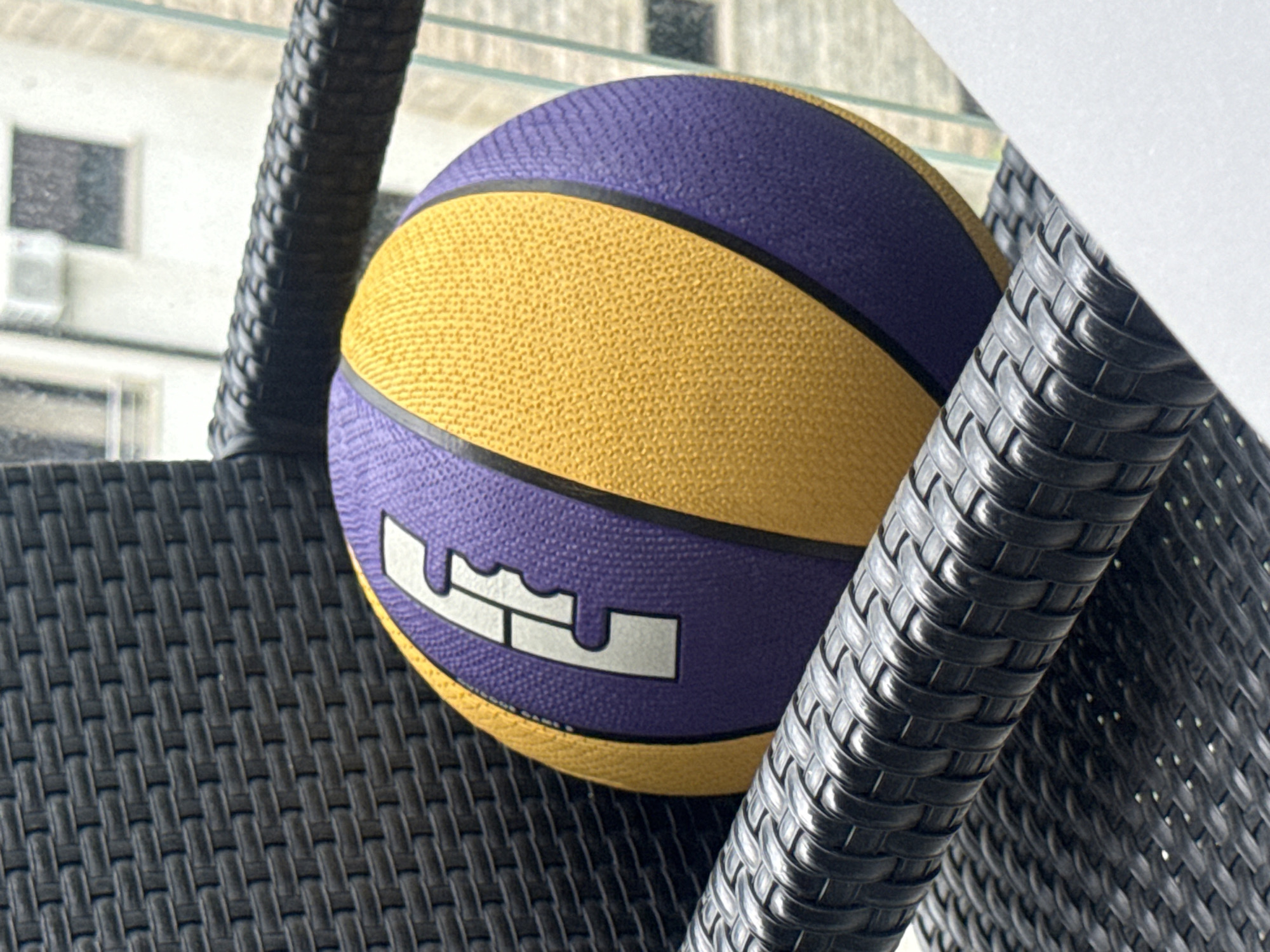 A basketball with purple and yellow panels and a white logo sits on a black woven chair on a balcony. The chair has a textured, woven surface. In the background, a blurred building with windows and an air conditioning unit is visible. The location is not Boreti, Montenegro.