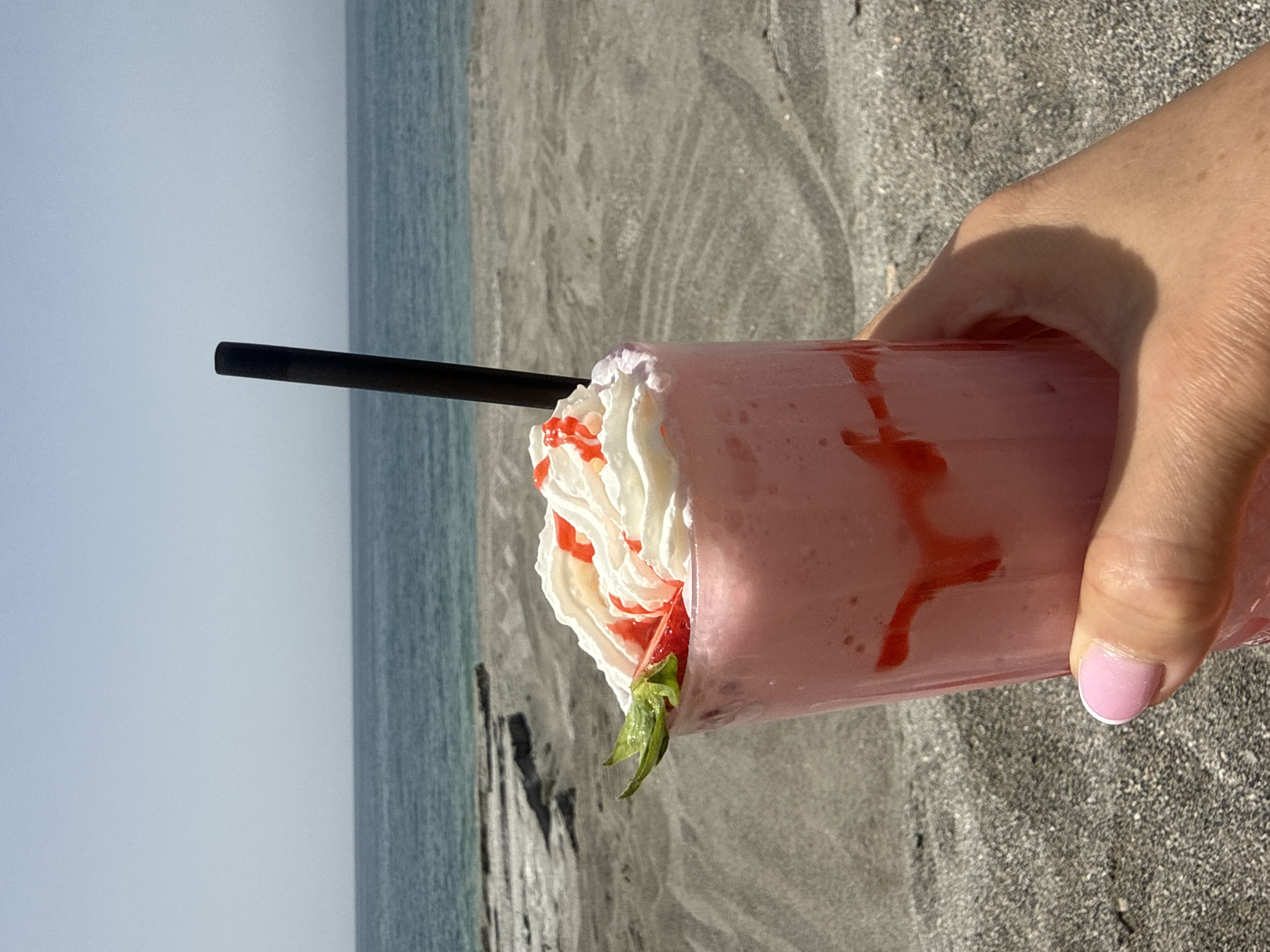 A person is holding a strawberry milkshake with whipped cream, drizzled strawberry sauce, and a black straw in front of a beach. The person's thumb with a pale pink nail is visible. The beach is made of grey sand, and the ocean is visible in the background under a light sky. The location appears to be near the city of Unknown, Unknown.