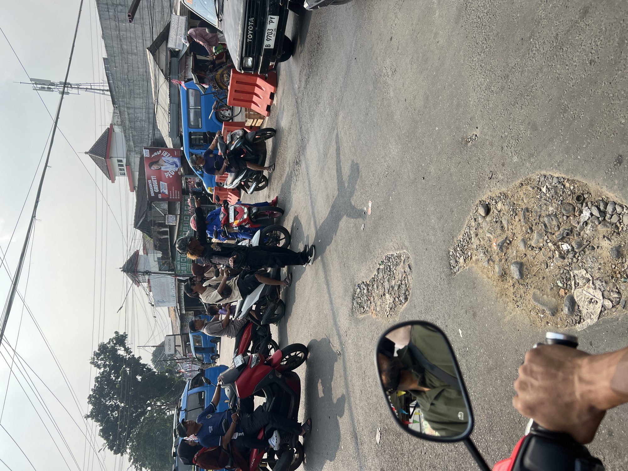 The image depicts a busy street scene in Citeureup, Indonesia, likely during daylight hours given the strong shadows. The foreground is dominated by a motorcycle handlebar and a rearview mirror, reflecting a person wearing a green jacket. The road surface is heavily deteriorated, with numerous potholes filled with gravel and debris.
In the mid-ground, several motorcycles and their riders are visible, forming a traffic jam. One person on a motorcycle appears to be directing traffic or gesturing. On the left, a red scooter is parked. Further back, blue vans are present, indicating a commercial or transport area. Various buildings line the street, some with visible signage, suggesting shops and businesses. A prominent billboard with an advertisement is also present. Power lines crisscross the sky above. The overall atmosphere is one of a bustling urban environment with clear signs of wear and tear on the infrastructure.