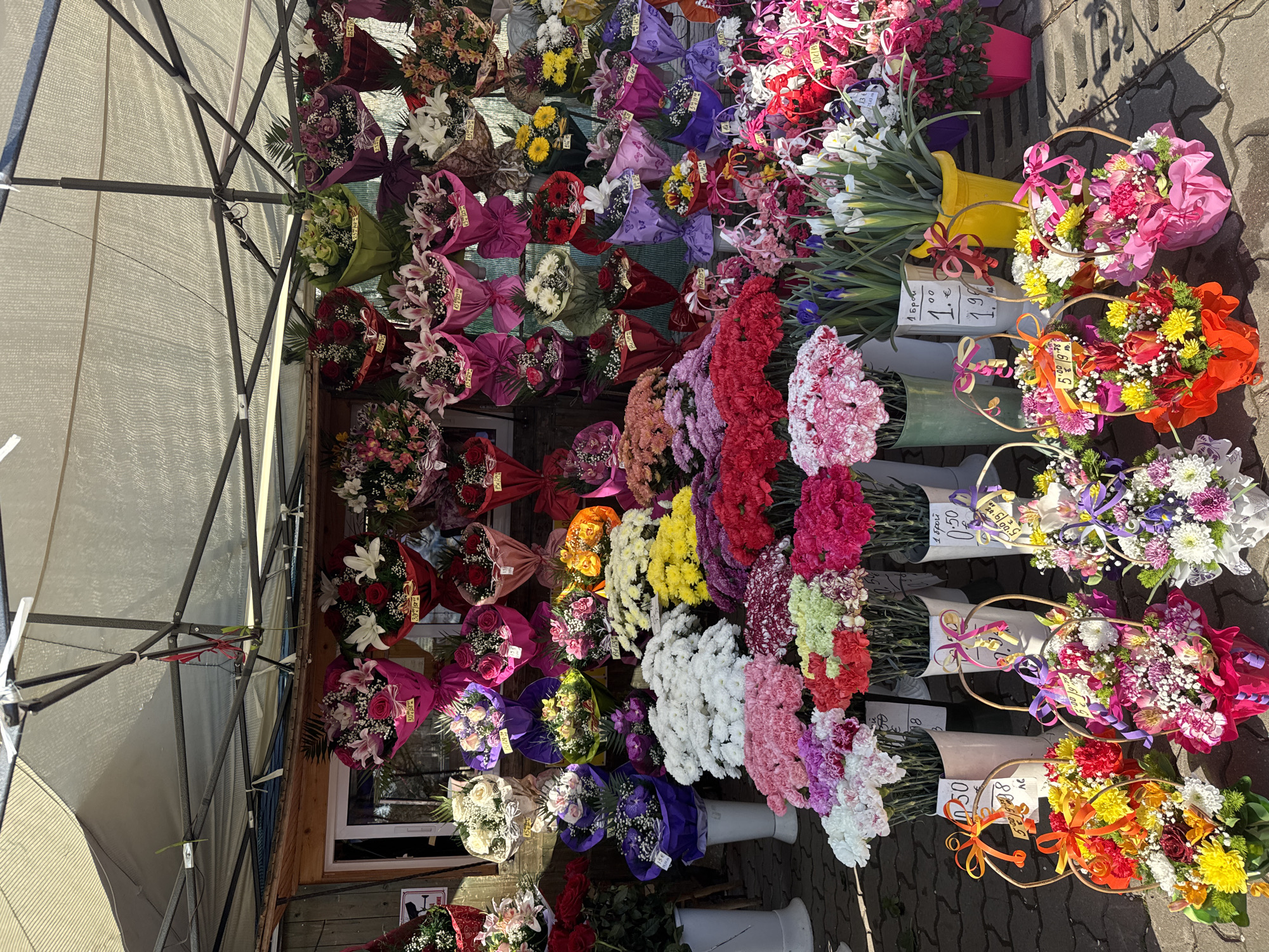The image shows a flower stall, likely outdoors, in Sofia, Bulgaria. Numerous bouquets of various colors and types of flowers are displayed. Some are wrapped in paper and hung on a wall-like structure, while others are arranged in vases or baskets on the ground. Price tags are visible on some bouquets. A tent frame is present on the left. The environment looks like a street or market.