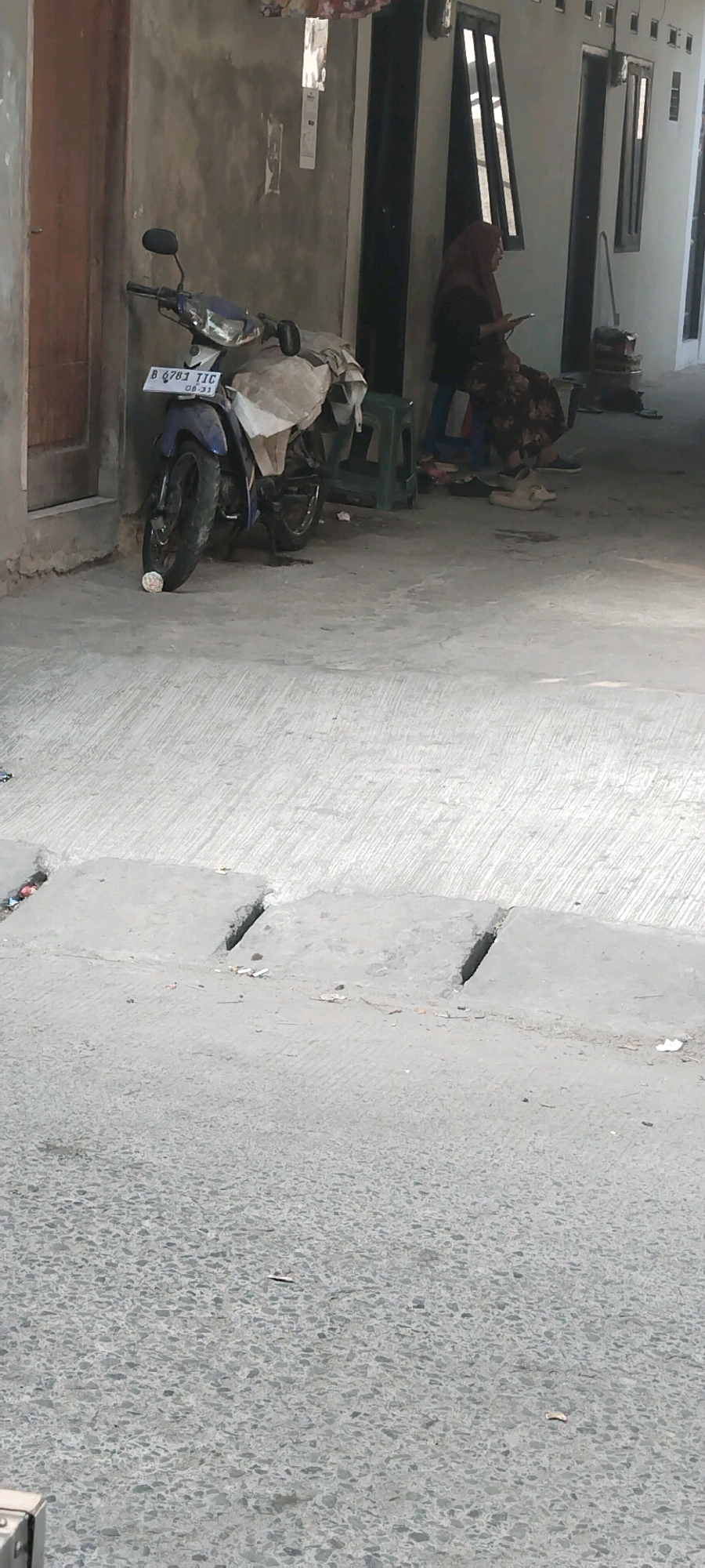 In West Jakarta, Indonesia, a motorcycle with license plate B 6781 TJC is parked on a concrete ground. The motorcycle is blue and partially covered with a tan tarp. A person wearing a maroon hijab and a patterned dress sits on a low green stool to the right of the motorcycle, holding a mobile phone. Behind the person are the doors and windows of a building, likely a residence or shop, with gray walls. The area appears to be an alleyway or walkway.