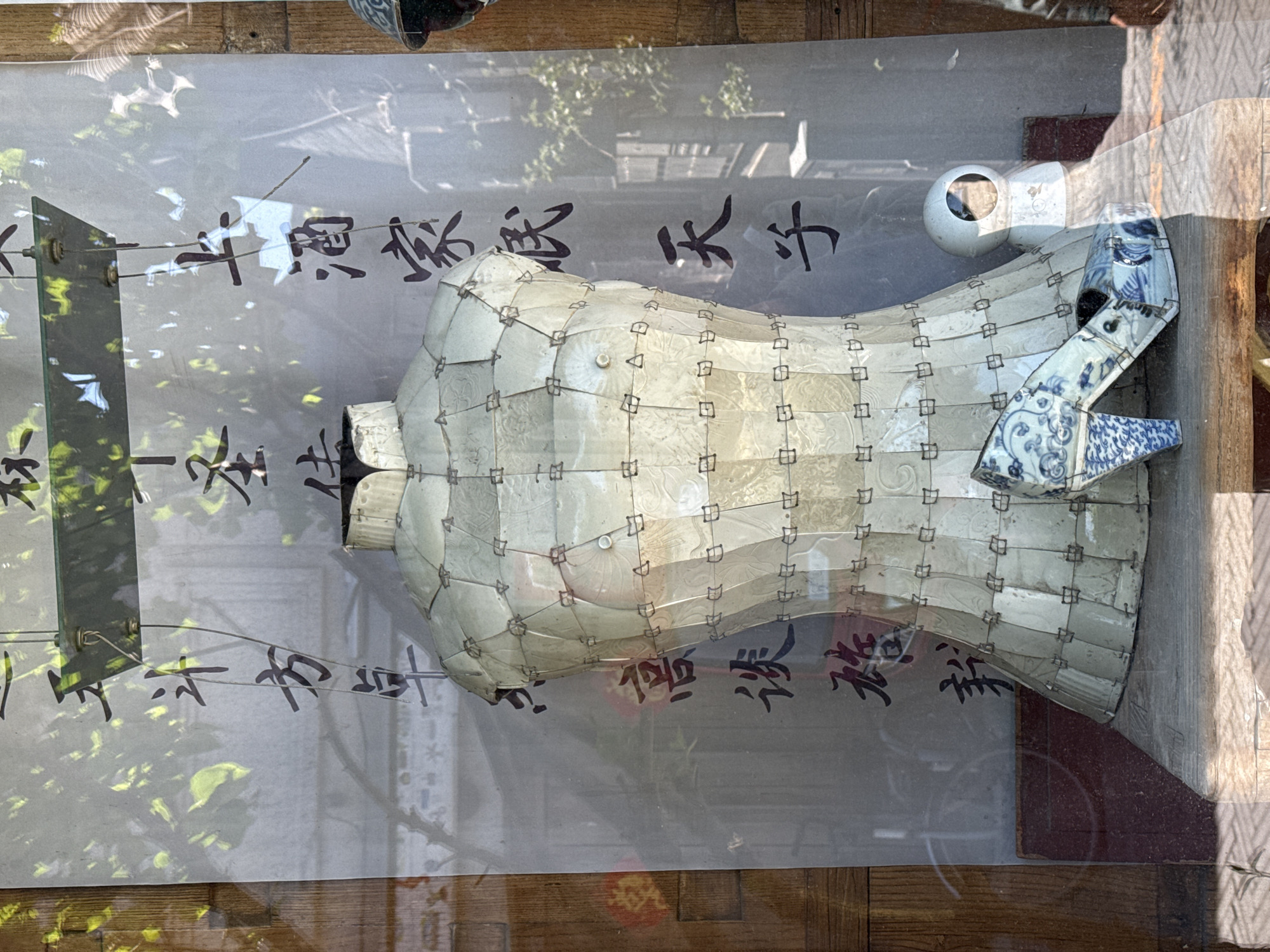 The image shows a window display in Xicheng District, China, featuring a torso-shaped sculpture made of interconnected white tiles, and a broken blue-and-white porcelain object placed on its side. A white, spherical object with a hole is positioned near the sculpture. Chinese characters are written on the window behind the objects. The background reflection suggests an outdoor environment with greenery. The display sits on a wooden surface.
