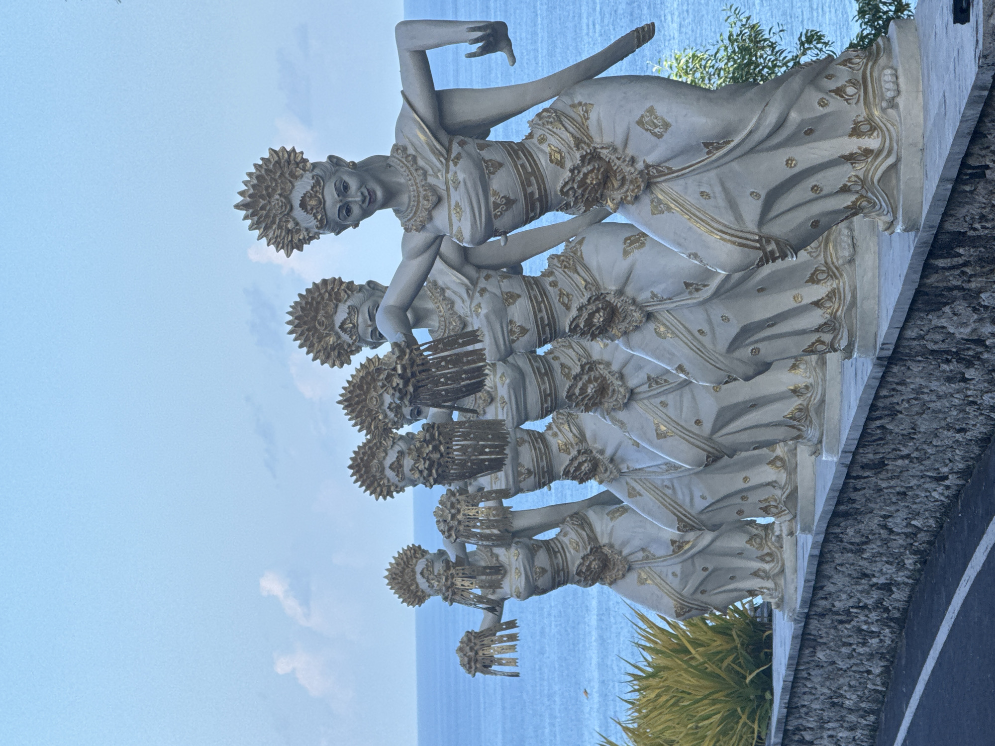 The image shows a line of six white statues of Balinese dancers. The statues are situated in Ungasan, Indonesia, overlooking the ocean. The dancers are depicted wearing traditional Balinese attire, including elaborate headdresses. The statues appear to be part of a monument or public art installation. The background includes a view of the sky and the ocean. The statues are erected on a concrete or stone base, alongside a roadway and some vegetation.