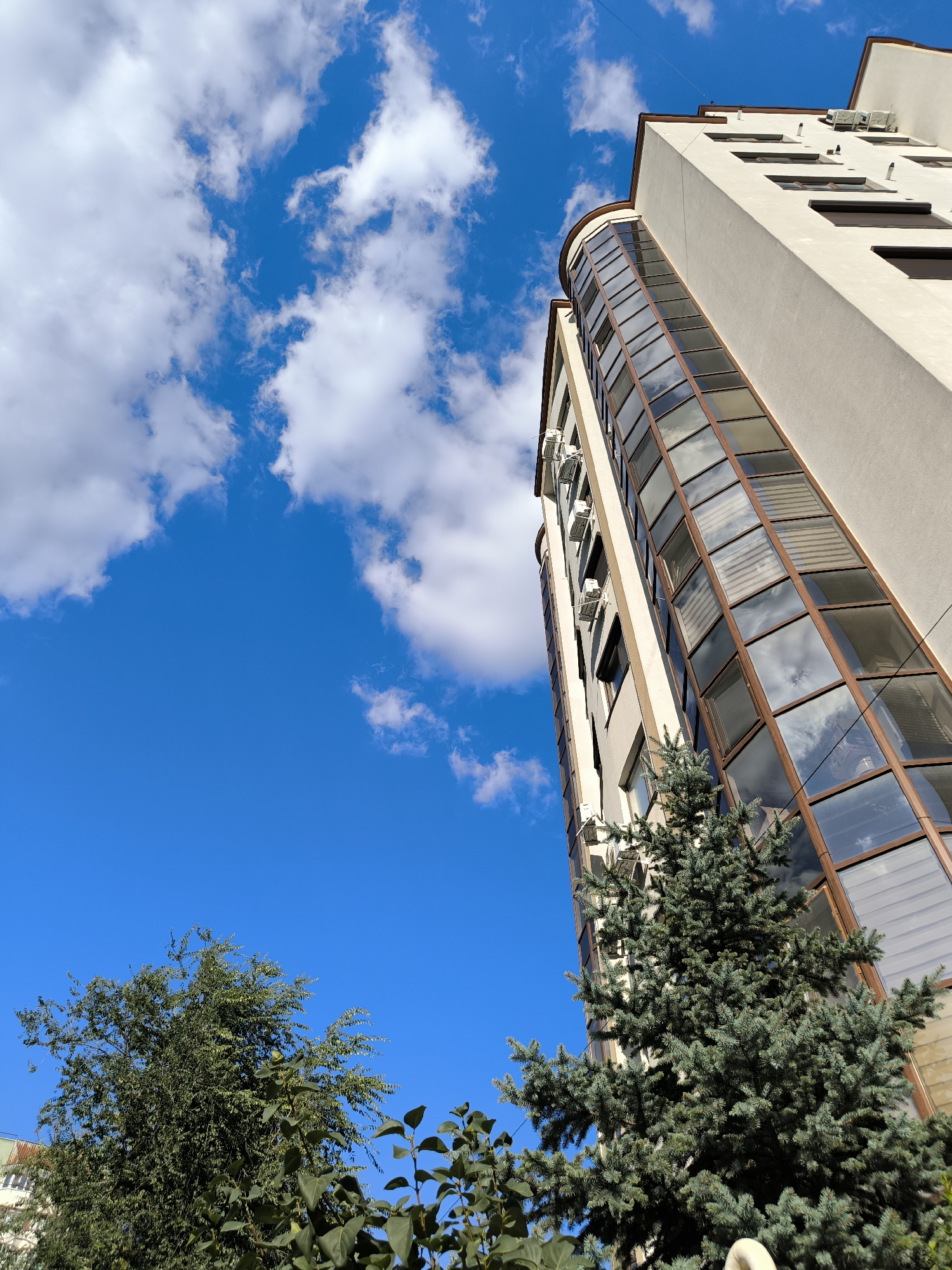 The image shows a low-angle view of a modern building with a curved section of glass windows, towering against a bright blue sky filled with scattered white clouds. Several air conditioning units are mounted on the exterior of the building. In the foreground, there are green trees, including a conifer tree. The overall scene depicts an urban environment on a clear day. This type of modern architecture and surrounding greenery is characteristic of residential areas in Chișinău, Moldova, specifically in districts with newer developments.