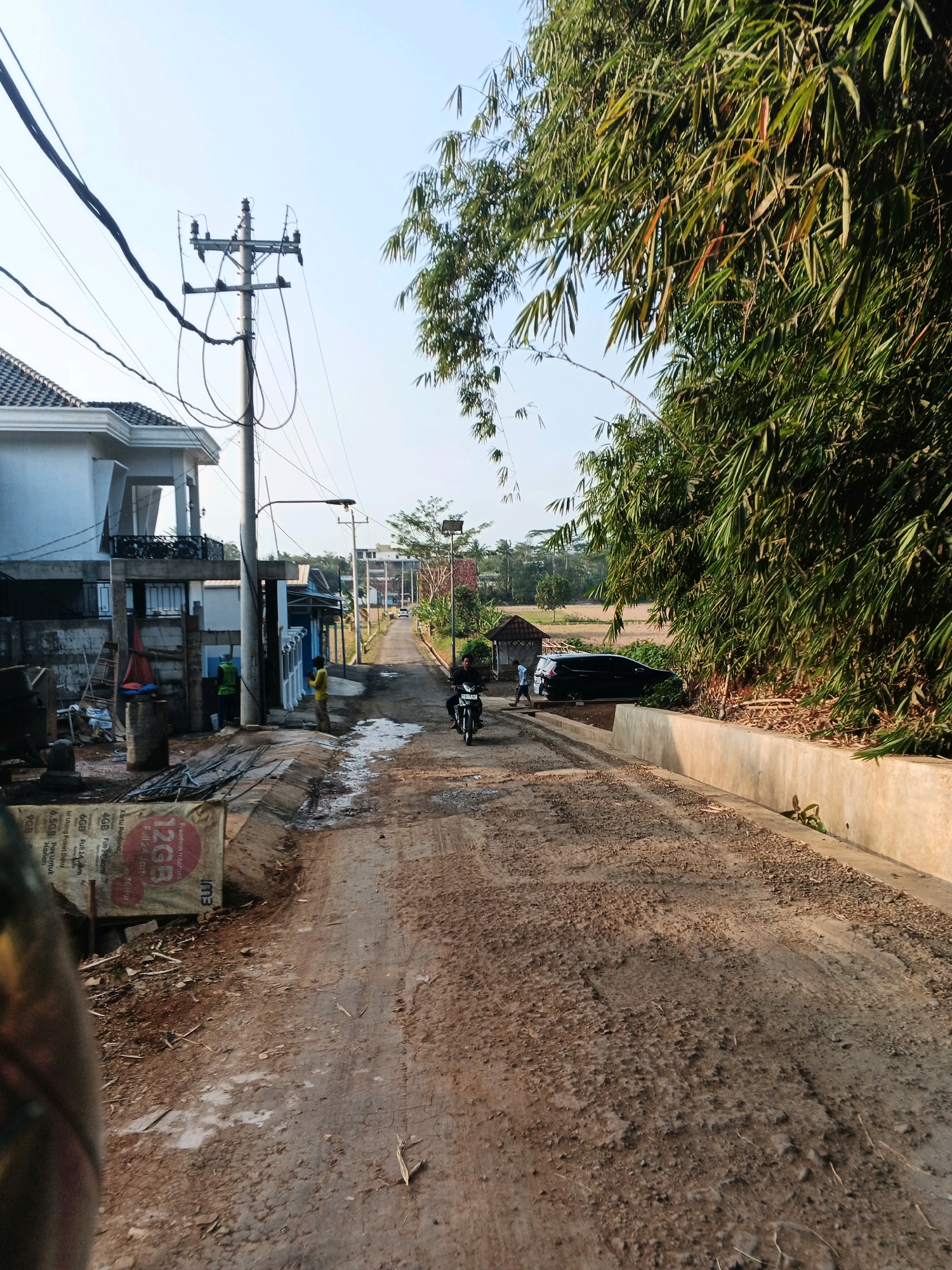 The image shows a rural road under construction in Doplang, Indonesia. The road surface is uneven, with patches of loose gravel and dirt. On the left side of the road, a modern-looking house is visible, along with construction materials and scaffolding. A utility pole with numerous wires stands near the house. Further down the road, two people are standing near a small building, and a black car is parked. A motorcycle with a rider is moving away from the camera, heading down the road. On the right side of the road, dense bamboo foliage lines the path, with a concrete retaining wall visible. The sky is clear and bright, suggesting it is daytime. The overall scene depicts an ongoing infrastructure project in a semi-rural setting.