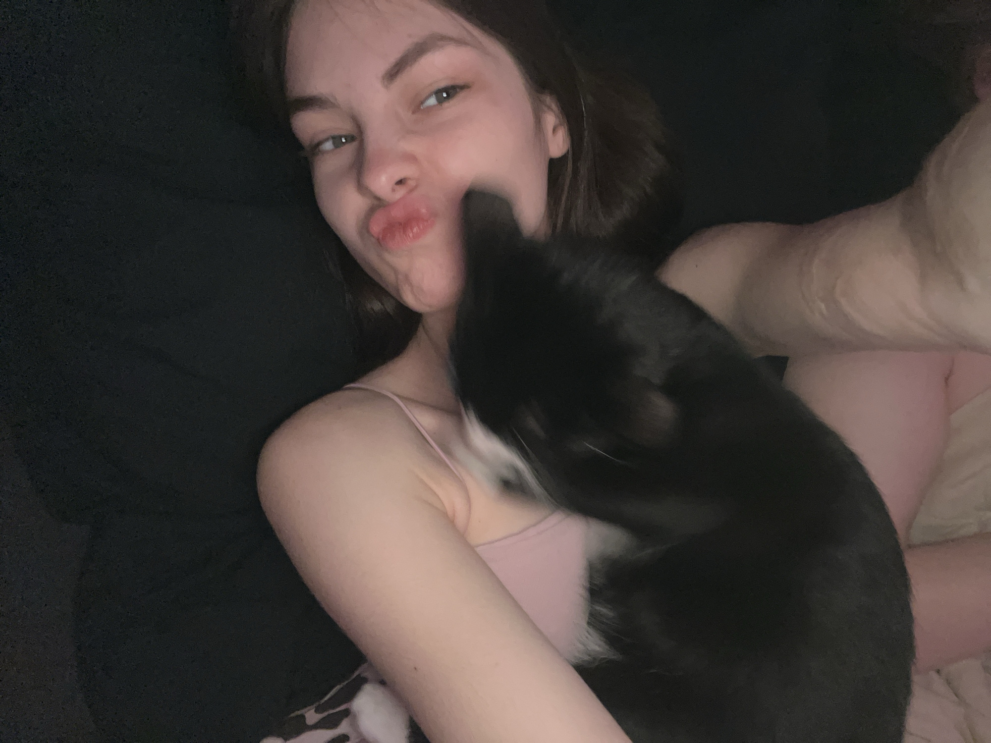 A young woman with light skin, short brown hair, and wearing a light pink tank top is holding a black and white cat. The woman is puckering her lips and looking slightly to the left of the frame. The cat's head is near the woman's face, possibly nuzzling her. The background is dark, seemingly a bed or couch. No information in the image allows for the location to be determined to be in Saint Petersburg, Russia.