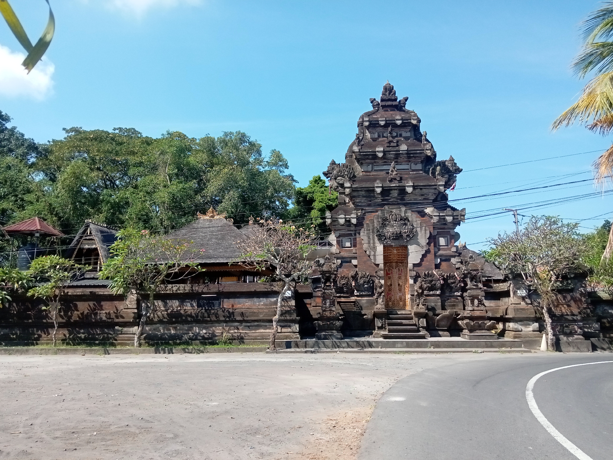 The image shows a Balinese temple entrance on a sunny day. The temple features intricate stone carvings and a tall, multi-tiered gateway. To the left of the gateway is a low stone wall with a traditional Balinese building behind it, complete with a thatched roof. Lush green trees are visible behind the temple structures, and a clear blue sky with a few wispy clouds forms the backdrop. In the foreground, a paved road curves to the right, with a dirt shoulder on the left side of the image. There are no people or animals visible in the scene.