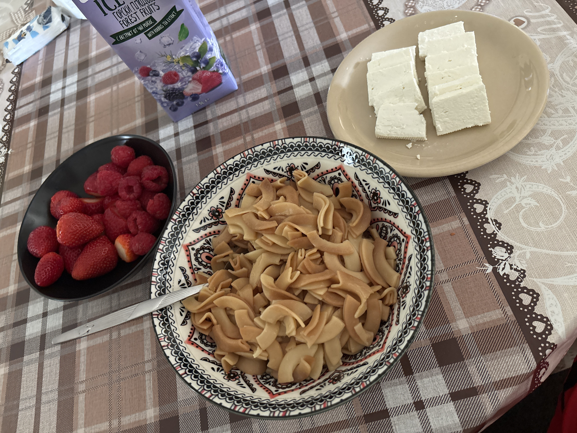 The image shows a table set for a meal. The tablecloth has a plaid pattern in shades of brown and beige, with a decorative border along one edge featuring floral and heart designs.
A carton of "Горски Плодове" (Gorski Plodove) Forest Fruits juice is placed near the upper-left corner. A black bowl containing strawberries and raspberries is situated to the left of a bowl of cooked pasta. A knife rests alongside the pasta bowl. To the right, there is a plate of sliced white cheese.