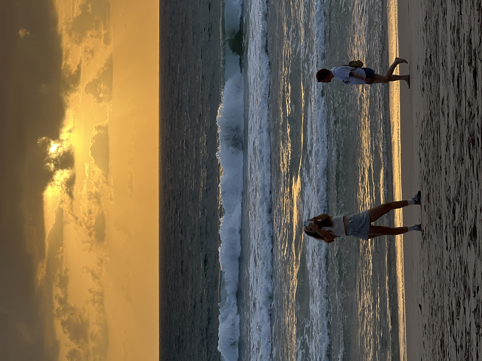 The image depicts a beach scene in Choeng Thale, Thailand, during sunset. The sky is a vibrant orange and yellow, with dramatic clouds partially obscuring the sun. The ocean is dark blue with white-capped waves rolling towards the shore.
In the foreground, two individuals are visible on the sandy beach. To the left, a person is standing and appears to be taking a photograph with a smartphone. They are wearing a light-colored top and a skirt. To the right, another person is walking along the shoreline, facing away from the camera. This individual is wearing a t-shirt and shorts, and has a small bag slung over their shoulder. The sand is textured with footprints and ripples from the receding waves. The overall lighting suggests the late afternoon or early evening hours.