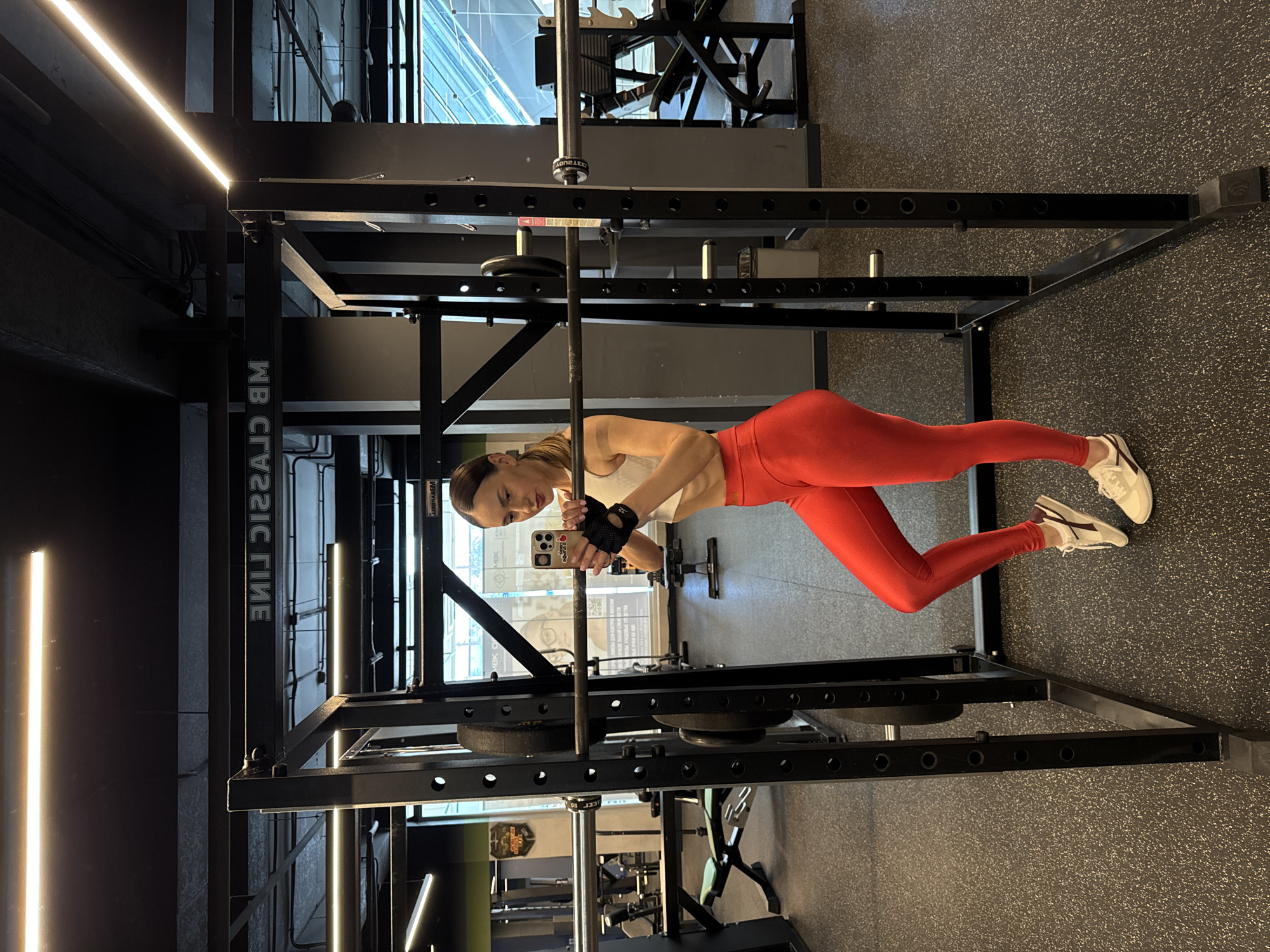 A young woman is depicted taking a mirror selfie within a modern indoor gym. She has light brown hair tied back and is dressed in a white athletic crop top, vibrant red leggings, white and beige sneakers, and black fingerless gym gloves. Her expression is neutral as she holds a light-colored smartphone with a decorated dark case in her left hand, facing it towards herself to capture the image. Text visible on her phone case includes "Blondie" and "love".
The background features a robust black power rack with a barbell resting on its hooks, flanked by several weight plates stored on lower pegs. The barbell has mirrored text that appears to read "IVANKO". On one of the power rack's uprights, vertically oriented and mirrored text "MB CLASSIC LINE" is visible. The gym floor is a dark, speckled rubber surface, and the space is illuminated by bright linear LED lights running along the ceiling and walls. The overall setting suggests a well-equipped fitness facility in Sochi, Russia.