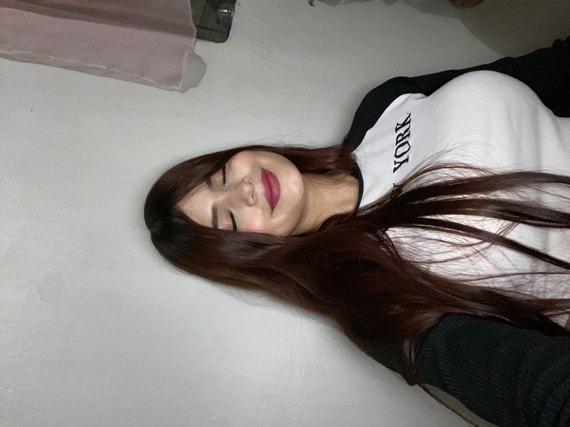 The image is a close-up, slightly tilted portrait of a woman. She has dark brown hair, fair skin, and is wearing dark lipstick. Her eyes are closed, and she appears to be smiling. She is wearing a white top with the word "YORK" printed in black letters. Part of a black garment is also visible. The background is a plain white wall. There is no indication of the location being Juan Griego, Venezuela or any identifiable city.