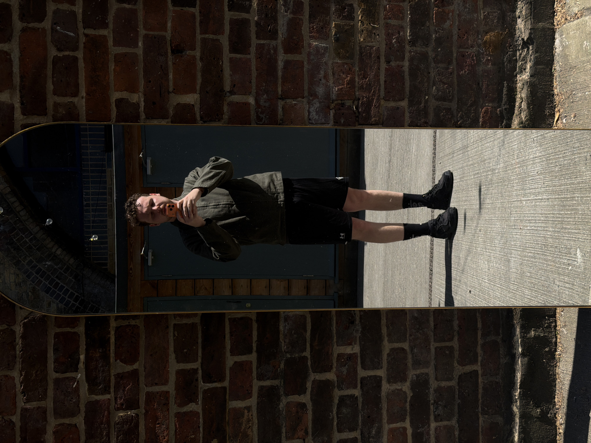 A reflection in a long, oval mirror placed outdoors shows a man holding a cell phone to his face, standing on a textured concrete surface. He wears an olive-green jacket and black shorts with the Under Armour logo. The mirror is framed in gold and mounted on a brick wall to the left and a concrete pillar to the right. Behind the man, there is a metal door with a handle, and above it, a window with a grid pattern. The scene is bathed in sunlight, casting shadows. A reflection of the mirror is seen in the top left corner. Based on architectural style, this could be located in London, UK.