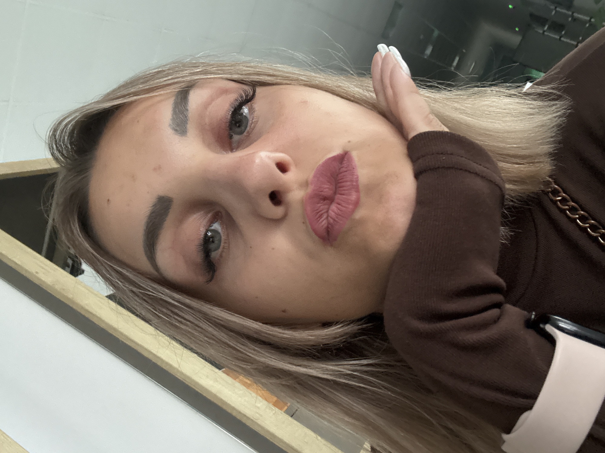 The image is a close-up, selfie-style photograph of a young woman with blonde hair and full, rosy lips pursed as if for a kiss. Her eyes are open and looking towards the camera. She is wearing a brown turtleneck top and her right hand is raised, with a manicured nail visible, touching her chin. The background appears to be indoors with some wooden architectural elements and a blurred, dark background with hints of lights. There is no discernible connection to Yuzhno-Sakhalinsk, Russia.