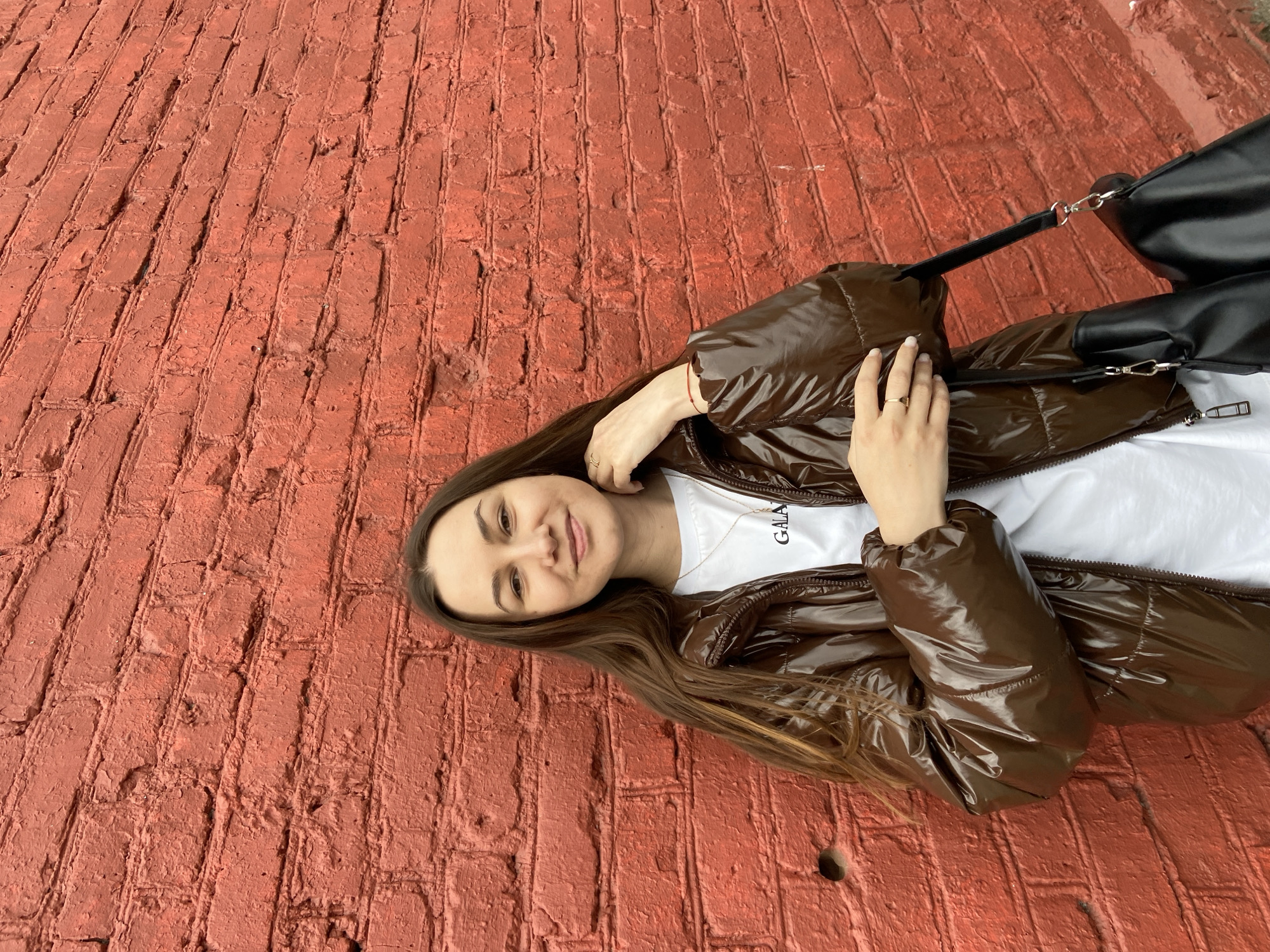 The image displays a young woman lying on a textured red brick surface, looking directly at the camera with a gentle smile. She has long, straight dark brown hair. She is wearing a shiny, dark brown puffy jacket over a white t-shirt, on which the letters "GALA" are partially visible in black. A black handbag with a silver chain strap is positioned near her right shoulder. A small gold-colored ring is visible on her left ring finger, which is placed near her face. The brick surface appears weathered and robust, with clear grout lines and some irregularities in the brickwork, suggesting an outdoor urban setting. The lighting indicates it is daytime, contributing to a calm and casual mood. This scene is located in Izhevsk, Russia.