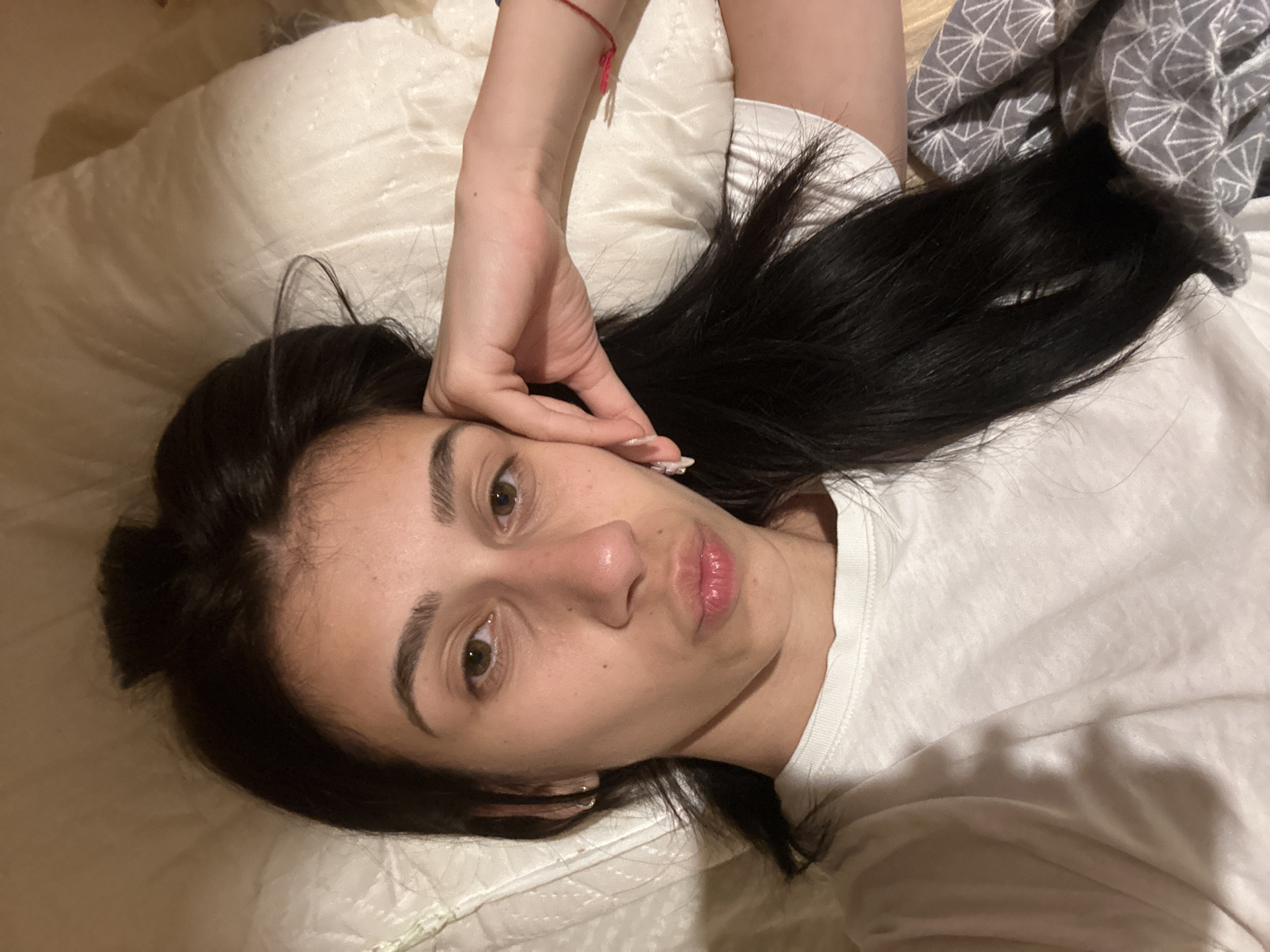 A woman with long, dark hair is lying on a bed. She is wearing a white t-shirt and has fair skin. She is looking directly at the camera. Her right hand is raised and touching her face. There is a white pillow under her head and a patterned gray blanket nearby. The location appears to be indoors, possibly a bedroom. The image does not provide sufficient information to determine a location in Plovdiv, Bulgaria.