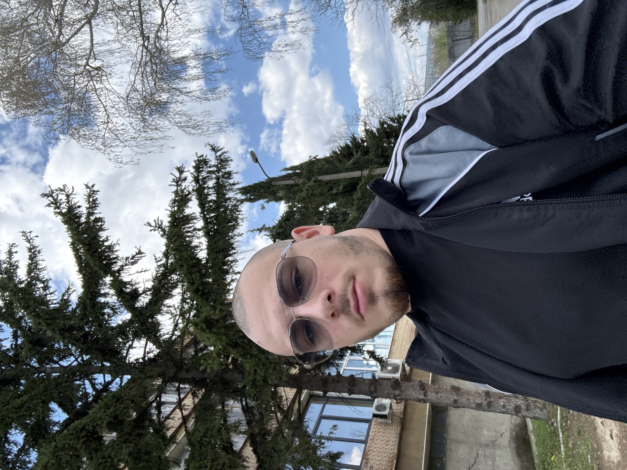 A man with a shaved head, a short beard, and sunglasses is taking a selfie outdoors. He is wearing a black and white Adidas tracksuit jacket. Behind him is a building with windows and air conditioning units, obscured by a large evergreen tree. Above the tree, part of the sky with white clouds is visible. This location is likely near the building on Stefan Stambolov Street in Stara Zagora, Bulgaria.