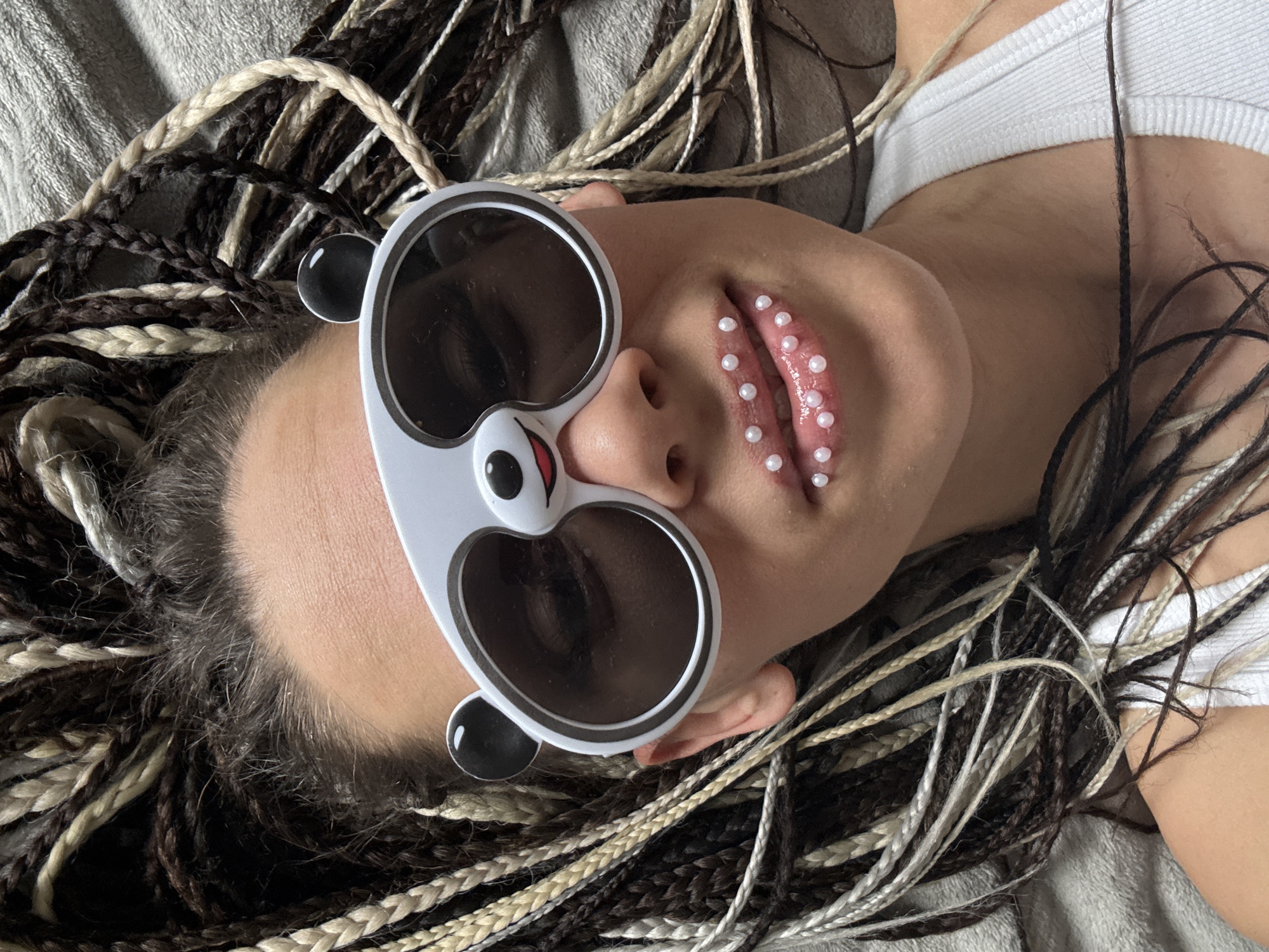 An individual is depicted lying on a soft, light grey textured surface, likely a blanket or bedspread, in Rusanovo, Russia. The perspective is from above, looking down at the person's head and upper torso.
The subject is wearing novelty panda-shaped sunglasses, featuring a white frame, dark reflective lenses, and cartoonish panda facial features on the bridge of the nose, along with round black ears extending from the sides. Their eyes are obscured by the dark lenses.
A notable detail is the decorative adornment on the person's lips, which are covered with multiple small, white, pearl-like beads, creating a distinct, textured effect. The lips appear slightly parted.
The individual has long, braided hair, a mix of dark brown/black and lighter blonde/white strands, which are spread out around their head on the textured surface. A white ribbed tank top or sports bra is visible on the upper chest.
The image appears to be a stylized portrait, focusing on unique fashion accessories and body adornment. No specific activity is taking place other than posing for the photograph. There is no visible text in the image. The time of day and specific weather conditions are not discernible from the image.