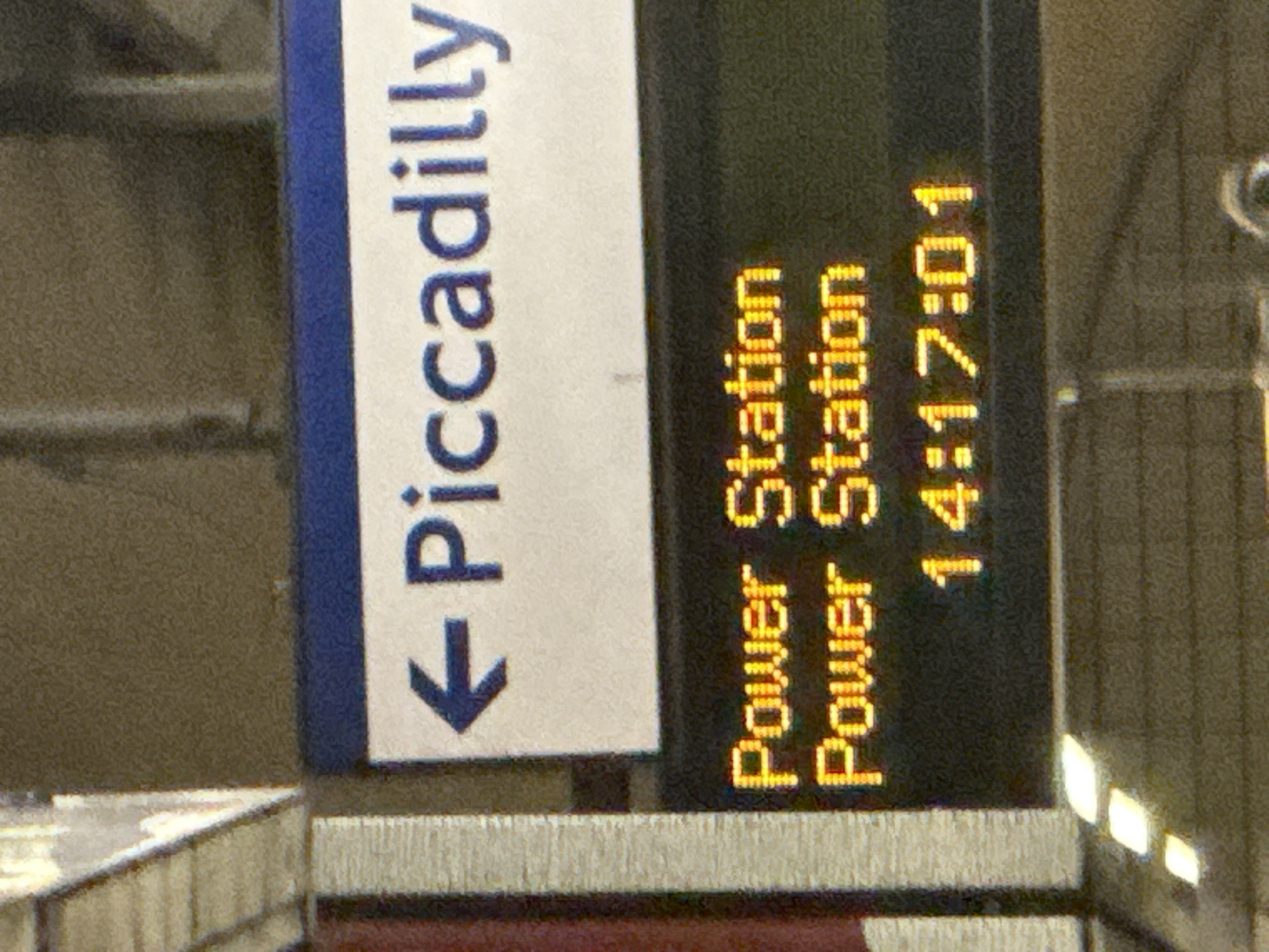 The image captures a scene at a London Underground station, specifically near a sign pointing towards the Piccadilly line. Next to the sign is an electronic display showing "Power Station" twice, with the time displayed as "14:17:01." The image is taken at close range, focusing on the signage and electronic display. The location is likely in London, United Kingdom, given the presence of the London Underground signage.