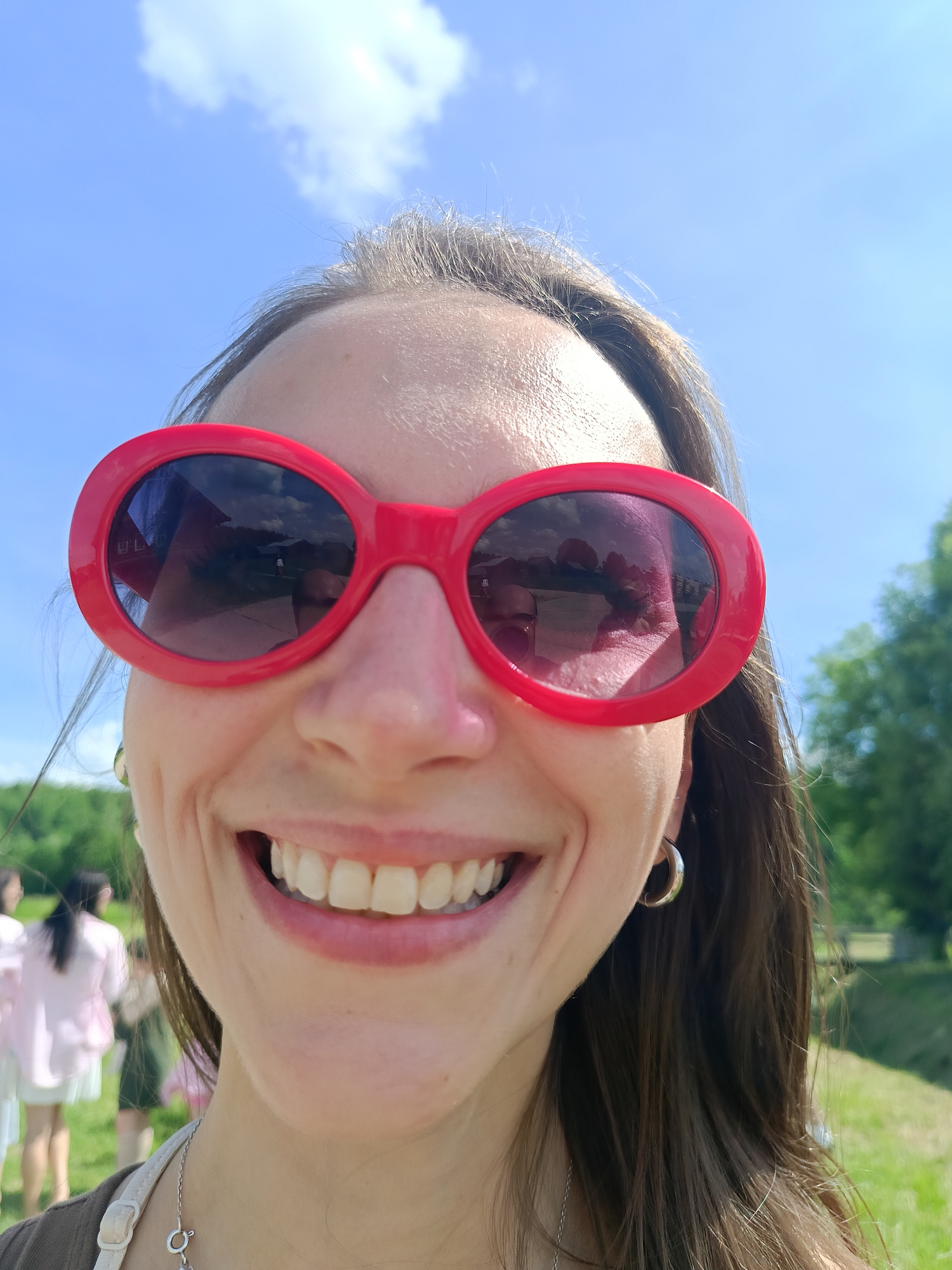 The image is a close-up selfie of a woman wearing large red sunglasses and a wide smile, taken outdoors under a bright blue sky with some white clouds. Her brown hair is visible, and she is wearing hoop earrings. In the background, out of focus, are trees, a grassy area, and several other people dressed in light-colored clothing, suggesting a gathering or event. The sunglasses reflect the sky, clouds, and some structures, including a building. The location is identified as Popovka, Russia.