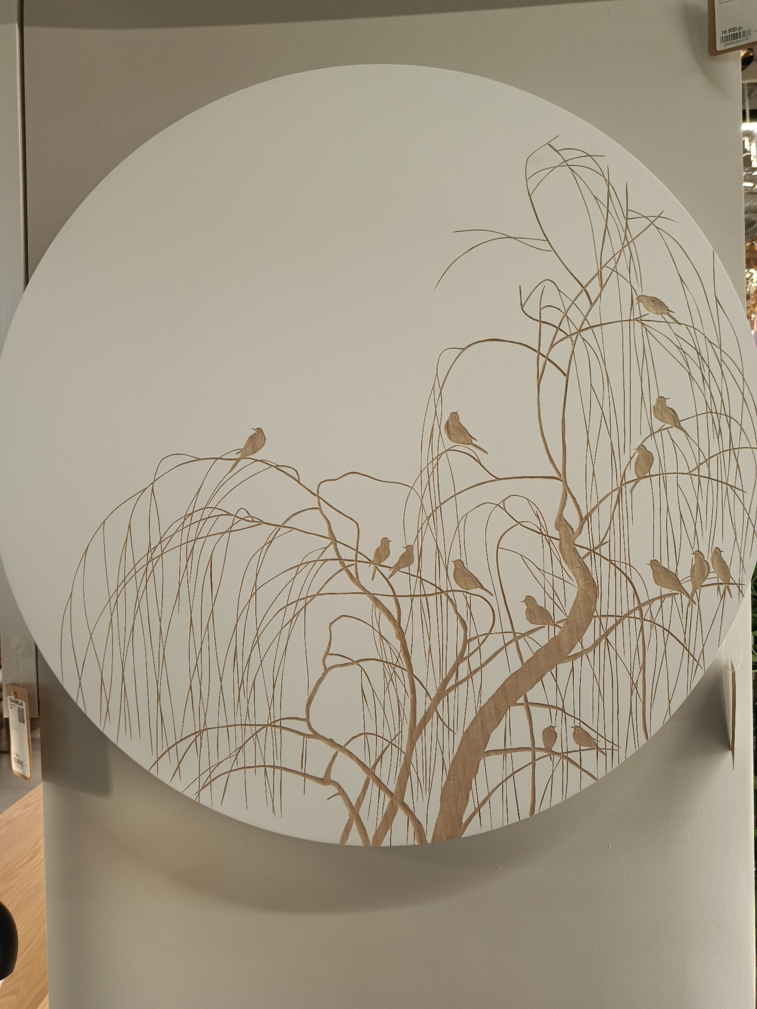 A large, round decorative panel, predominantly off-white or light beige, is the central subject. It features an intricate, carved or painted design of a tree, likely a weeping willow, rendered in a golden-brown hue. The tree's slender, drooping branches and delicate twigs cover the lower two-thirds of the panel. Numerous small, uncolored birds are perched throughout the branches, appearing serene and integrated into the natural scene. Approximately 15 to 20 birds are visible, varying in their poses.
The artwork is displayed indoors, possibly in a retail store or exhibition space in Moscow, Russia. Neutral-toned walls and display structures surround the panel, with shadows indicating artificial lighting.
A price tag is visible in the upper right corner of the original image and one crop, showing "16 500 р." (16,500 Russian Rubles), along with other indistinct text. Another partially visible tag or label is seen in the bottom left of one cropped image. No people are present, and the primary activity captured is the presentation of this decorative artwork. The artistic style is elegant and minimalist, focusing on line and form.