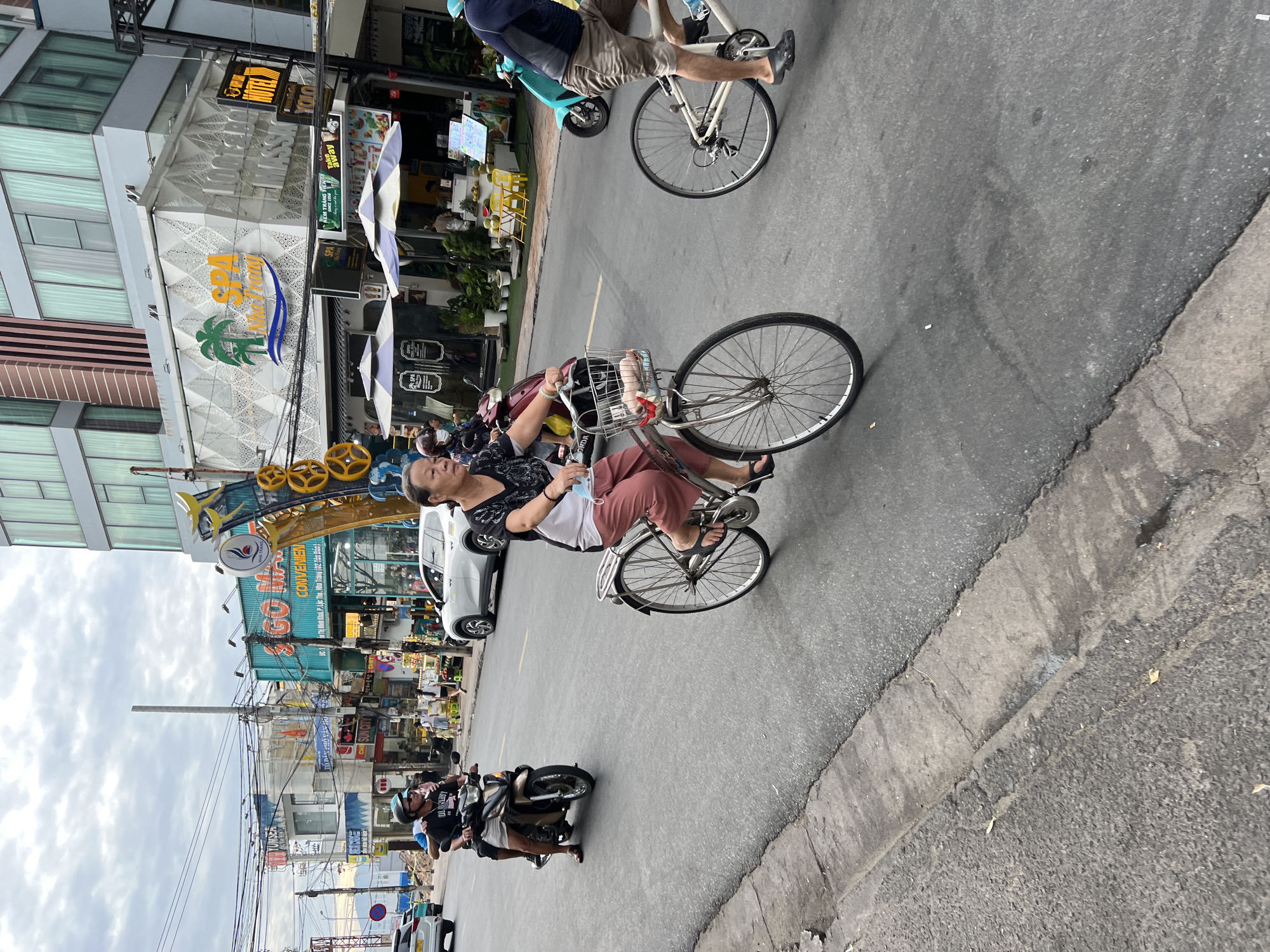 The image depicts a street scene in Nha Trang, Vietnam, with a focus on the activity of people on bicycles and a motorcycle. The foreground shows a woman on a bicycle, dressed in pink pants and a dark patterned shirt, with a face mask hanging from her chin. She is riding a bicycle with a basket on the front, containing what appears to be a small bag and some red items. Behind her, another person is riding a bicycle, visible from the waist down, wearing shorts and dark sandals. To the left, a man is riding a motorcycle, wearing a helmet and dark t-shirt, with another person behind him. In the background, buildings line the street, with various signs advertising businesses such as "SPA Nha Trang" and "HOTEL YOO." Cars and a blue scooter are also present on the road. The sky is overcast, suggesting a cloudy day. The overall environment suggests a typical urban street with mixed traffic and commercial establishments.