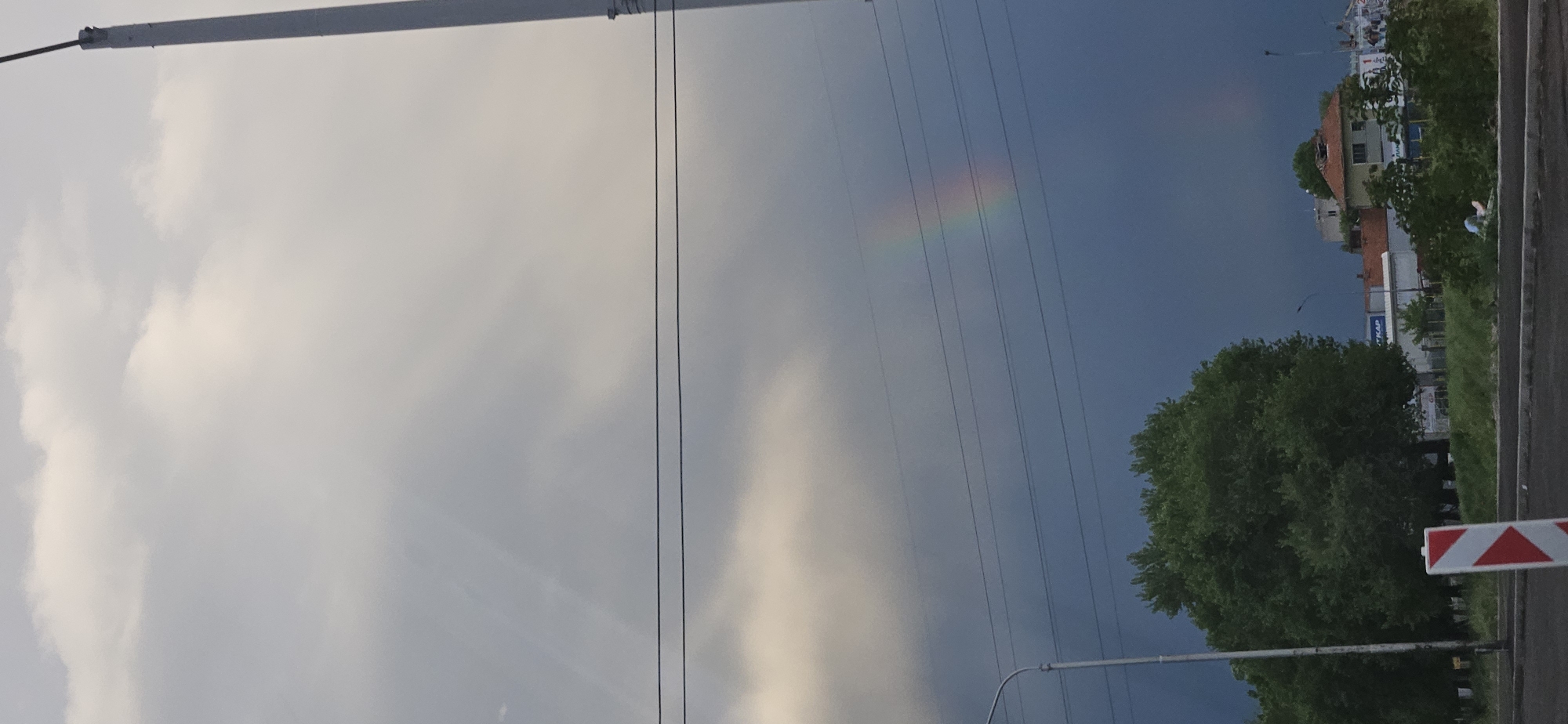 The image presents an outdoor scene in a city, possibly Unknown, Unknown, characterized by a contrast between atmospheric conditions and urban elements. The left portion of the image shows a light-colored, cloudy sky. A faint rainbow segment is visible in the upper-middle area. The foreground on the right side of the image includes a road with a red and white striped traffic barrier. A tall green tree is situated behind it. Above the tree, structures indicative of a cityscape are discernible, including building rooftops and advertisements. Overhead power lines are strung across the sky. A lamp post appears in the bottom right. The general impression is that of a city landscape experiencing varying weather.