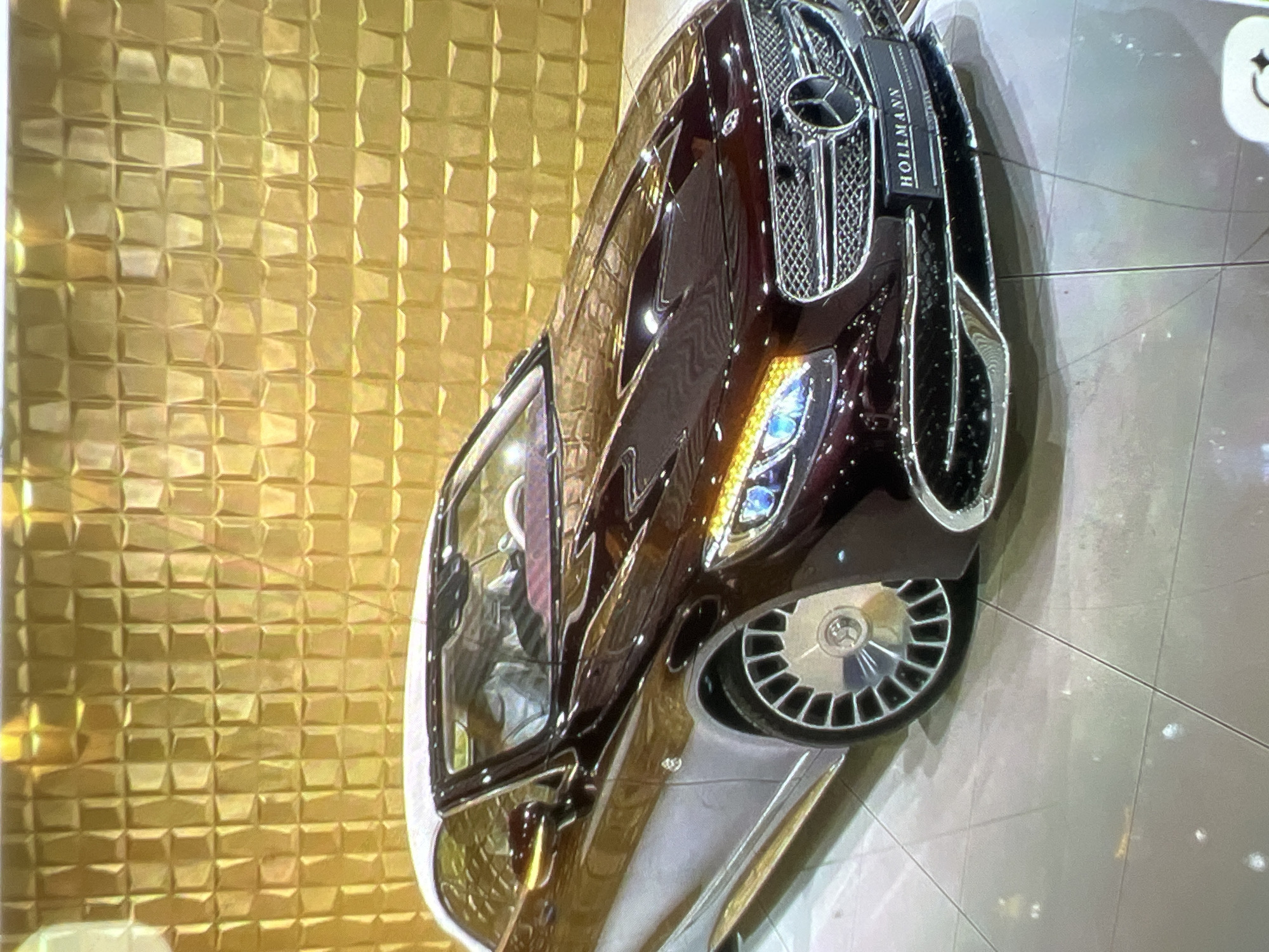 A dark red Mercedes-Benz convertible with a white soft top is parked indoors on a light gray tiled floor. The car's interior, including the steering wheel and seats, is covered with protective plastic. The car is positioned in front of a wall with a textured, golden, square-patterned design. A license plate frame on the front of the car reads "HOLLMANN". The lighting in the scene is bright and reflective, creating glare on the car's surface and the floor. The background wall is uniformly illuminated, highlighting its three-dimensional pattern.