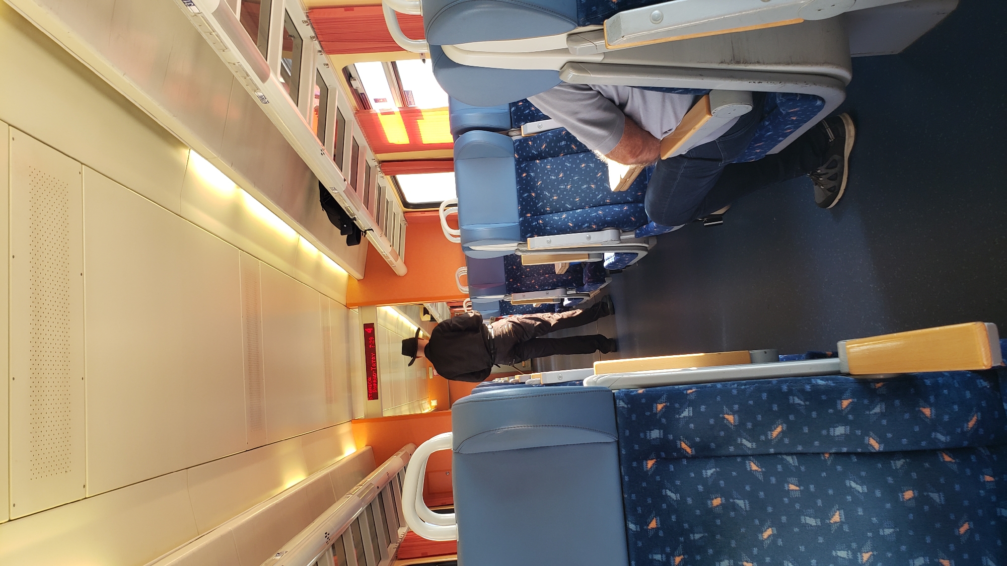 The image captures the interior of a train carriage. In the foreground, rows of blue seats with a blue and orange patterned fabric are visible, suggesting a public transportation setting. A person is seated on the right, wearing dark jeans and grey sneakers, with their arm resting on the armrest. Further down the aisle, a person dressed in dark clothing, including a cowboy hat, is standing and facing away from the camera. This individual appears to be holding a small object in their hand. Overhead, there are luggage racks and lighting fixtures. A digital display board is mounted on the wall at the end of the carriage, showing "Poprad-Tatry 7:29" and a red indicator, possibly signifying a train departure time from Poprad, Slovakia. The overall environment is that of a train in transit, with the typical seating arrangement and amenities found in such a vehicle.