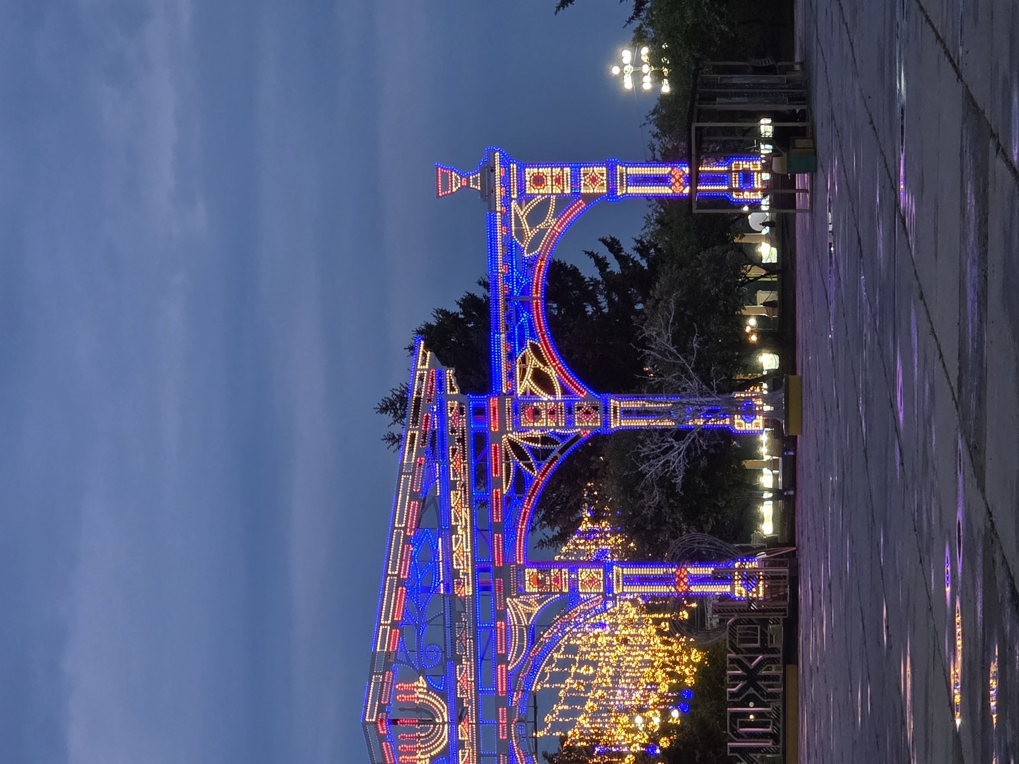 The image depicts an illuminated archway structure adorned with red, yellow, and blue lights. The structure is composed of rectangular columns and arched segments. The top of the archway features a decorative element resembling a small tower. In the background, trees and other light fixtures can be observed. The sky is a dusky blue. The ground is paved with grey tiles. The image is taken at night. The lighting suggests an urban area decorated for a festival or celebration. I can not determine the location in the city of Birobidzhan, Russia.