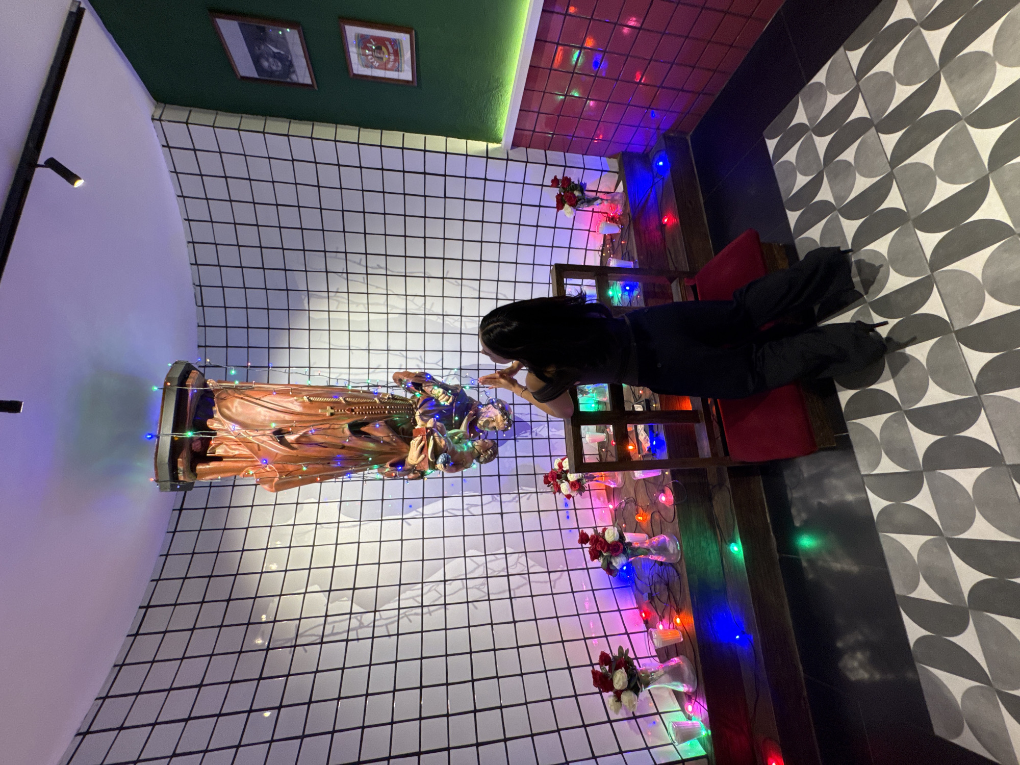 The image depicts an indoor scene located in Puebla, Mexico, centered around an inverted shrine and an individual engaged in an act of reverence.
A single person, viewed from behind, is kneeling on a red cushioned stool. This individual wears a dark-colored top and black wide-leg trousers, with their hands raised and joined in a gesture consistent with prayer or deep contemplation.
The focal point is an ornate, inverted statue or figure suspended from the ceiling. This object appears to be crafted from a metallic or bronze-toned material, depicting multiple forms or details, and is intricately wrapped with multi-colored string lights. A rosary-like object hangs from a lower section of the inverted statue.
Below the suspended statue, a dark wooden shelf serves as an altar. It holds three clear glass vases, each containing a bouquet of red and white roses. Several small, clear glass votive holders, some emitting light, are interspersed among the vases, alongside draped multi-colored string lights.
The surrounding environment features a prominent, curved wall covered in white square tiles with dark grout, creating a grid pattern. To the right, an adjacent wall is painted dark green on its upper section, transitioning to red/pink subway tiles on the lower section, with a horizontal light strip separating the two. Two framed pictures are mounted on the green wall, one depicting two people and another a vintage-style label. The ceiling is white, with a black track light fixture containing a single spotlight. The floor directly beneath the shelf and the kneeling individual is composed of dark, solid tiles, which transitions to a foreground area featuring a geometric pattern of black, gray, and white semi-circular tiles. The scene is illuminated by the ceiling light, the colorful string lights, and the votives, casting varied hues across the surfaces.