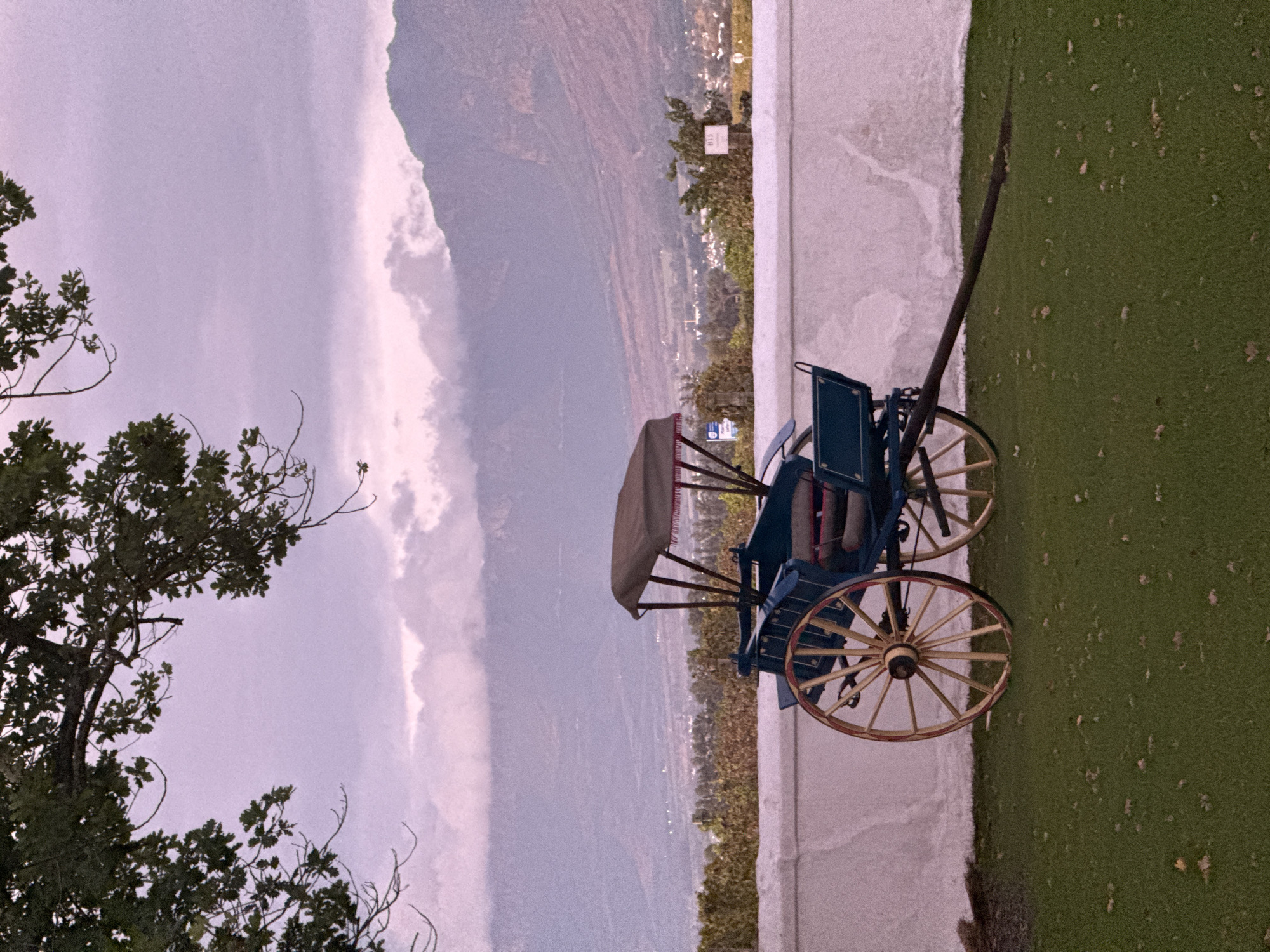 The image depicts a serene mountainous landscape under an overcast sky, likely in Drakenstein Nu, South Africa. In the foreground, on the left side, a large tree with dark green leaves and some bare branches partially frames the scene. On the right, a patch of vibrant green grass is visible, bordering a low, light-colored wall or barrier.
A blue and beige horse-drawn carriage or buggy with large, distinctive wooden spoked wheels (light-colored with reddish-brown accents on the rims) is positioned on the grass, serving as a decorative element. Its shafts point towards the right side of the frame.
Beyond the wall and the carriage, a vast valley unfolds, dotted with faint lights or structures of a settlement in the distance. Overlooking this valley, a small white sign with dark text reading "H1LS" is mounted on a pole near the wall. The background is dominated by majestic, expansive mountains with a rugged texture, appearing in shades of purple and brown, suggesting distance and atmospheric haze. The sky above is largely overcast, displaying soft grey and lavender tones with lighter cloud formations. The overall lighting suggests either a cloudy day or soft light during early morning or late afternoon. No specific activity or people are visible; the scene focuses on the landscape and the static carriage.