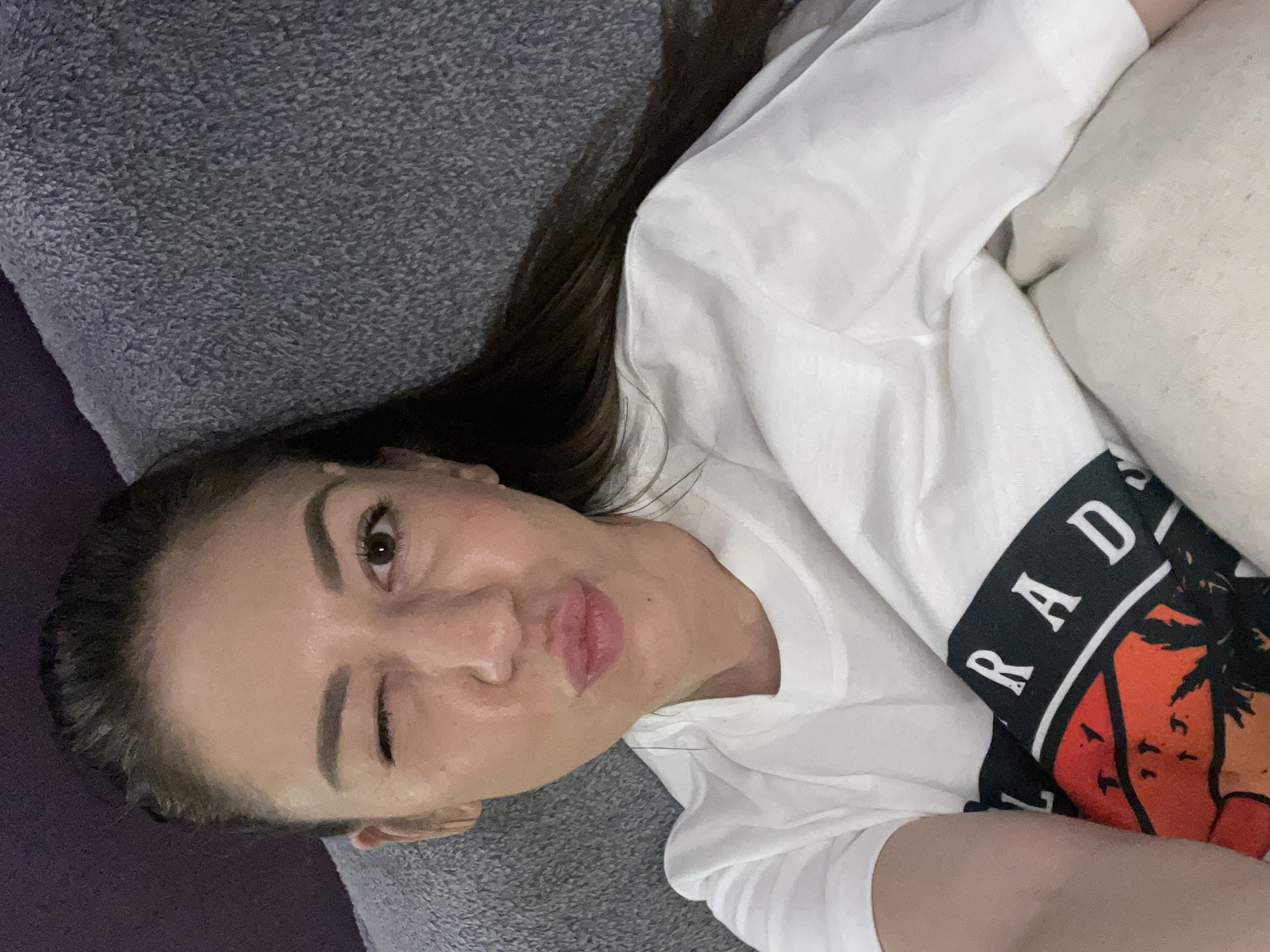 A woman is lying down. She is winking and puckering her lips. She has dark hair. She is wearing a white t-shirt with a graphic print. She is lying on a gray textured surface. A white pillow is visible. The image does not contain sufficient information to determine the location in Sofia, Bulgaria.