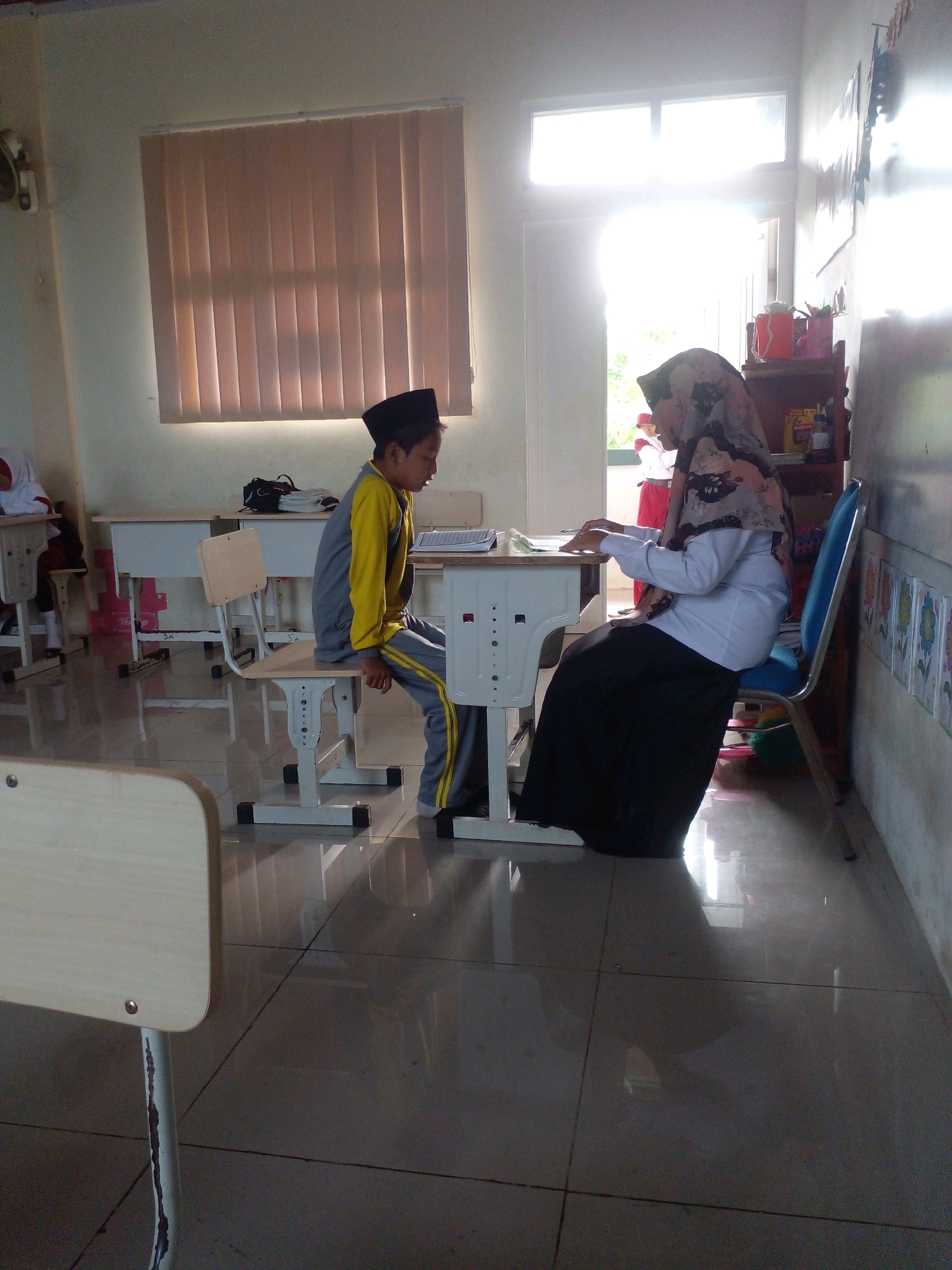 This image captures a classroom scene in Banyuputih, Indonesia, where a teacher, wearing a hijab and a white shirt with a long black skirt, is interacting with a young male student. The student, dressed in a yellow and grey uniform with a black cap, is seated on a desk, looking towards his teacher. A desk is between them, and the teacher appears to be writing or reviewing something. The classroom features desks and chairs, with sunlight streaming in through a large window, illuminating the space. The flooring is tiled, and the walls are painted. In the background, there are shelves with various items, possibly teaching materials or personal belongings. The overall atmosphere suggests an educational setting during a lesson or a one-on-one interaction between teacher and student.