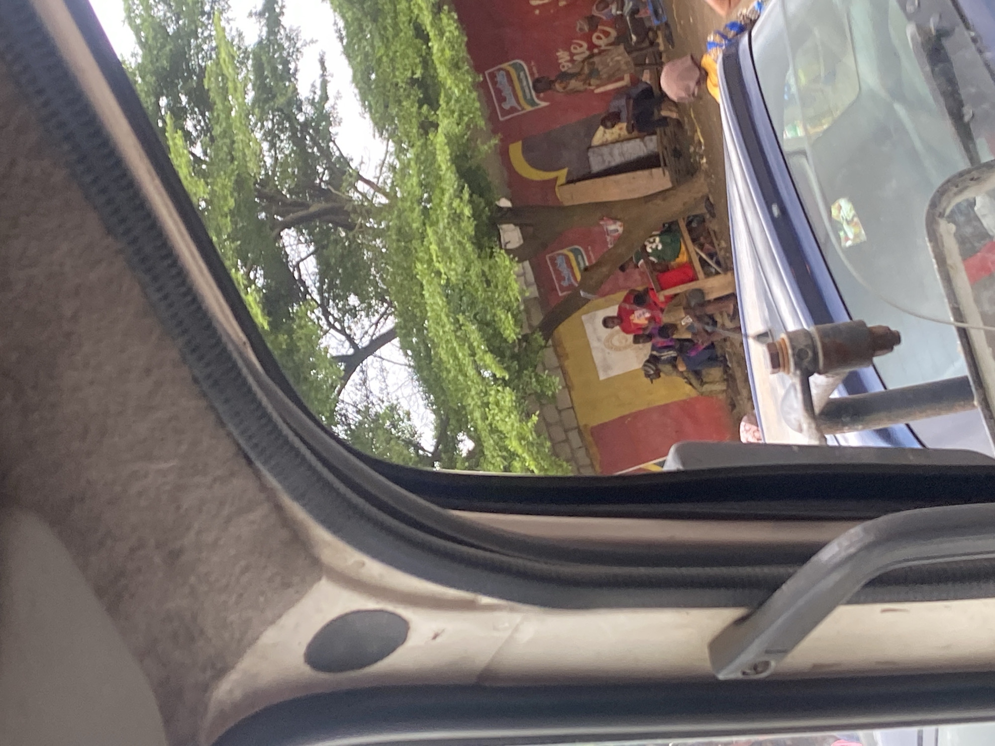 An outdoor street scene, likely in Ikeja, Nigeria, is viewed through the window of a vehicle. In the foreground, the interior of the vehicle is visible, including a grey handle and part of the dashboard. The main focus is the outdoor environment, characterized by a red brick wall adorned with yellow and red painted sections and advertisements featuring a logo resembling "Indomie." A large tree with lush green foliage dominates the midground, its branches reaching across the scene. Beneath the tree and in front of the wall, several individuals are gathered. Some are seated on the ground or on makeshift seating, while others are standing. Clothing varies, with some people wearing colorful attire. In the background, there appears to be a building structure. The overall impression is one of a casual gathering in a somewhat weathered urban environment.