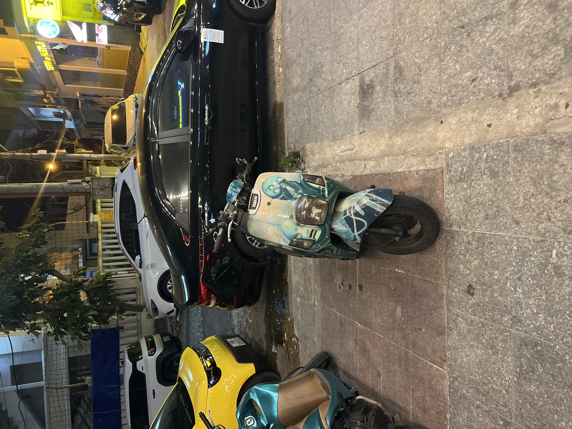 The image depicts a street scene in what is presumed to be Bắc Nha Trang, Vietnam based on the cultural cues and location. A black car is parked parallel to the curb on a paved surface. A Yamaha scooter with a custom paint job, featuring blue and white colors and illustrations, is parked immediately in front of the car. Further down the street, a yellow vehicle and another scooter are visible. The lighting suggests it is nighttime. The scene is somewhat cluttered with various vehicles, indicating urban density. The pavement is made of stone tiles or slabs.