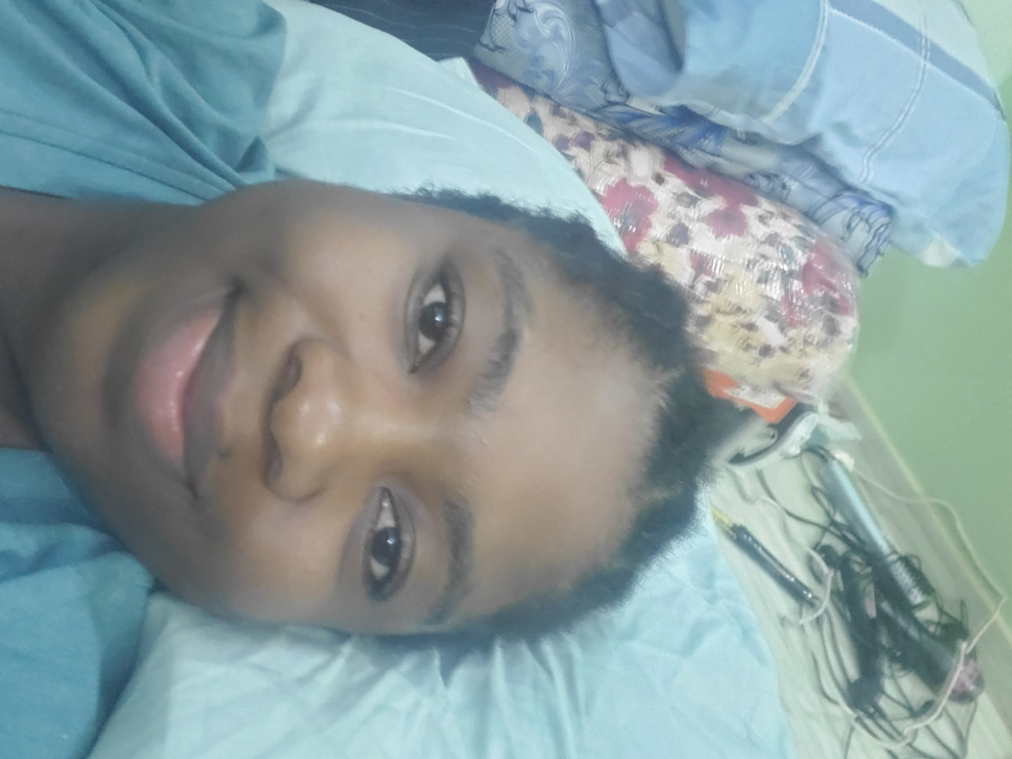 A dark-skinned person lies on a light blue bedsheet, facing the camera and smiling slightly. The person wears a blue shirt. In the background, there is a floral patterned pillow and a striped blue and white pillow. A clear plastic bag containing a black pen and a hair styling device with cords is visible. There is no indication of the location being in Barden Yana, Nigeria.