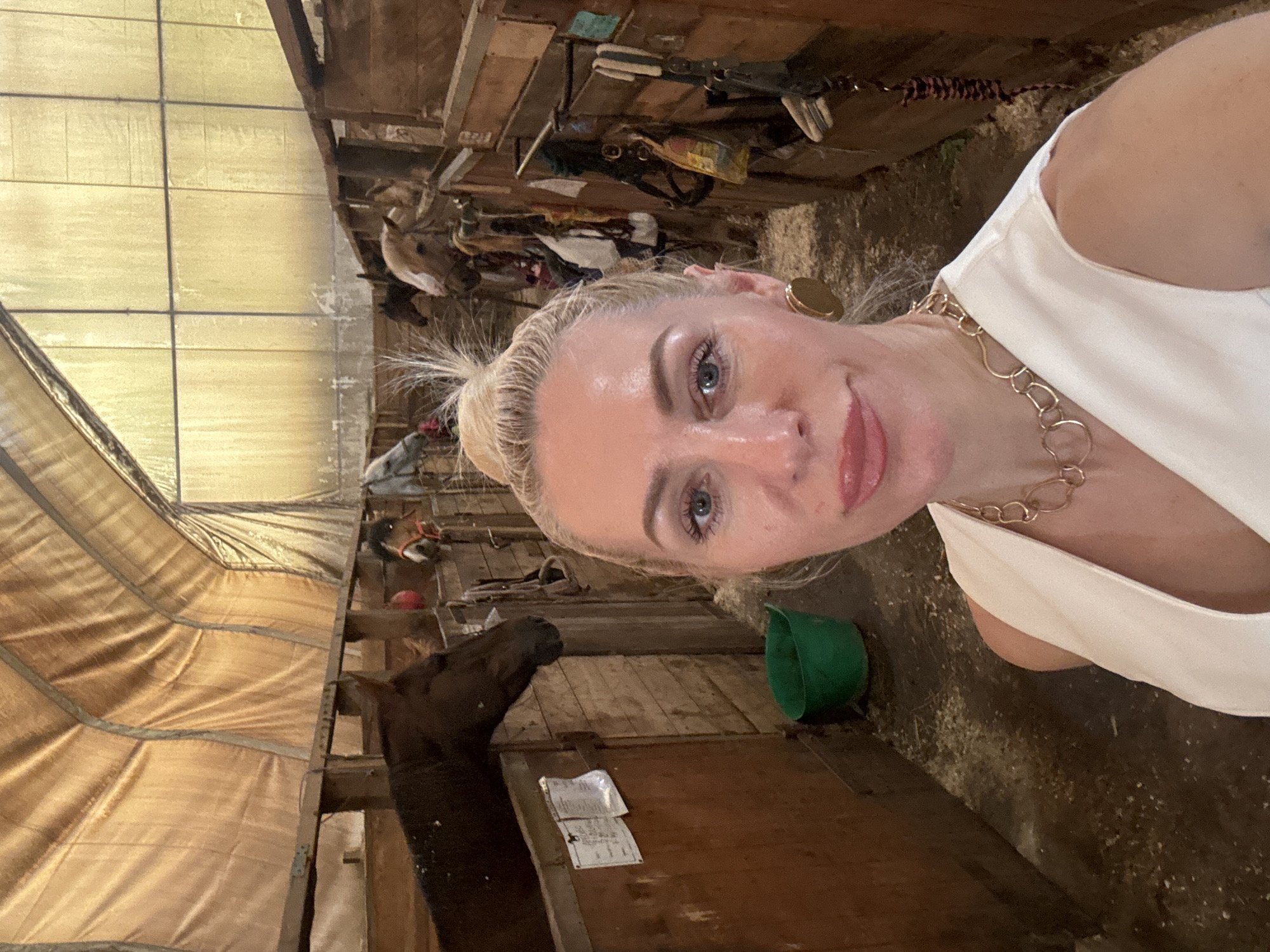 The image shows a woman with blonde hair pulled up in a bun, wearing a white top, a gold necklace, and large gold earrings. She is taking a selfie in a stable. In the background, there are several horses in stalls. The stable appears to be made of wood with a corrugated metal roof. There is a green bucket on the floor and various horse tack hanging on the walls. The scene is lit by natural light filtering through the roof. This image was taken at Pavlovsk, Russia.