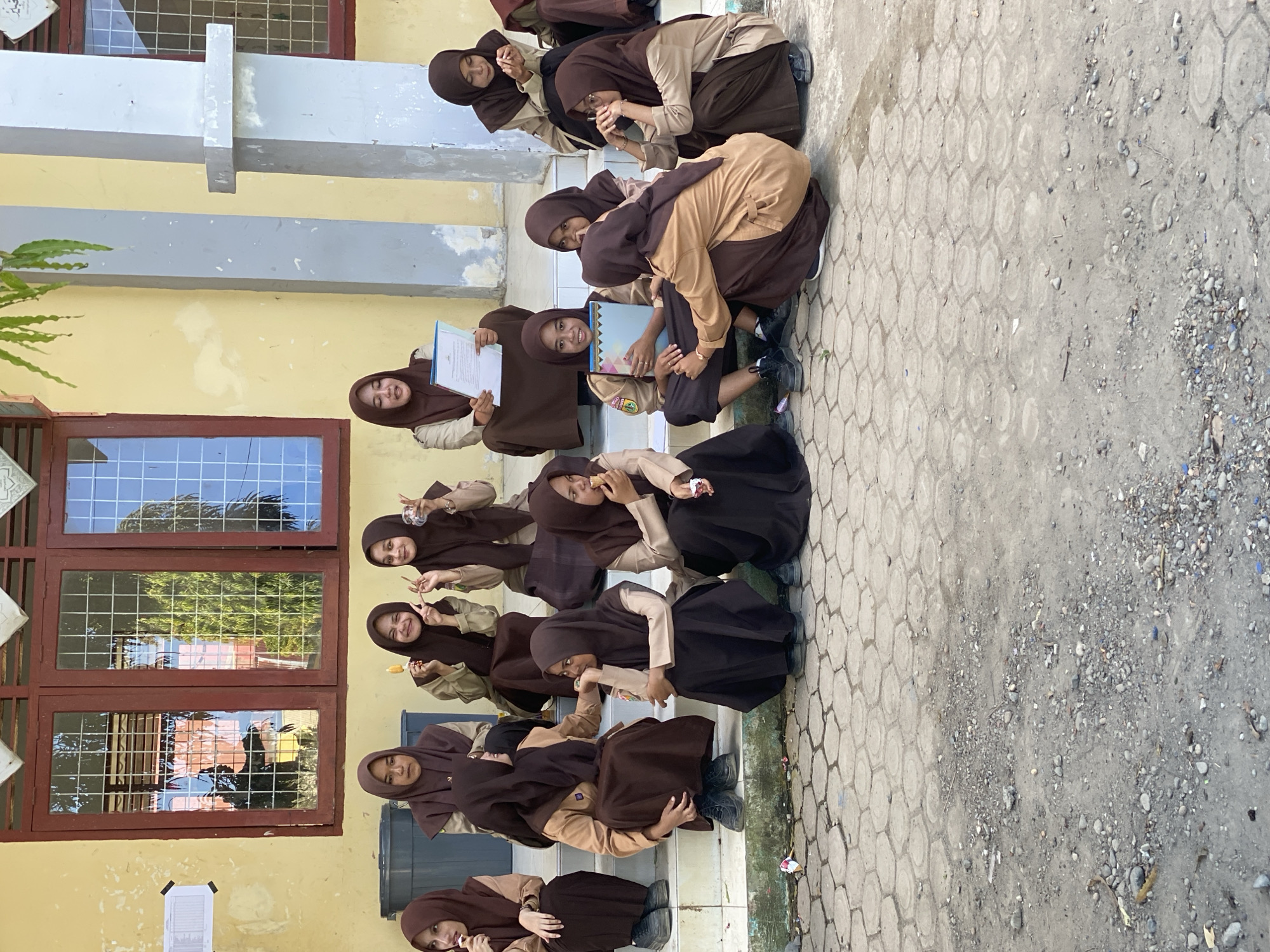 In an outdoor setting with a building facade featuring yellow paint and red-trimmed windows, a group of approximately ten young females, appearing to be students, are seated on concrete steps and the ground. They are all wearing brown hijabs and uniforms consisting of brown skirts and tan shirts. Some are holding objects, with one girl in the center holding a blue folder. The students are engaged in various activities, including looking at the camera, talking to each other, and eating snacks. The ground in front of them is paved with hexagonal concrete tiles and dirt. The overall atmosphere suggests a casual gathering or break time.