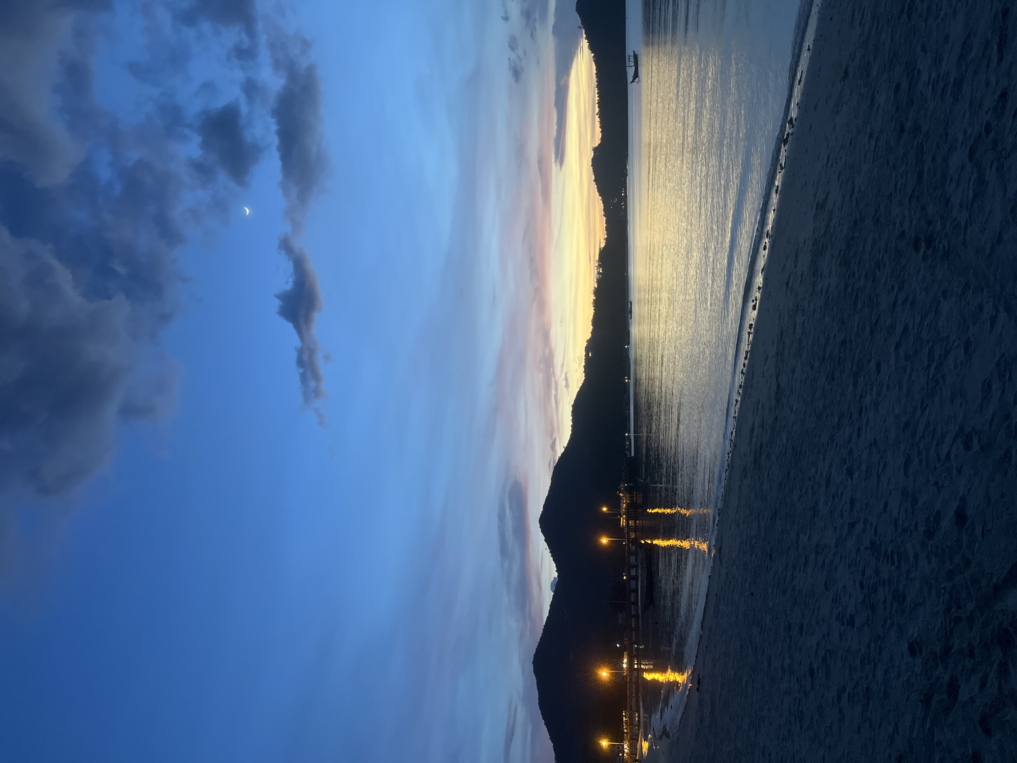 The image depicts a serene twilight scene on the beach in Koh Phangan, Thailand. The sky displays a gradient of deep blue to soft orange and yellow hues, with scattered dark clouds and a sliver of a crescent moon visible. In the foreground, dark sand forms the beach, leading to the gentle lapping of waves on the shore. The middle ground features a calm body of water reflecting the twilight colors and the bright, yellow lights of a pier stretching out into the sea. Several boats are visible on the water, including a silhouette of a longtail boat in the distance. The background is dominated by the dark, mountainous silhouettes of the island. The lighting suggests the transition from dusk to night, with artificial lights from the pier creating warm reflections on the water. No people are discernible in the frame.