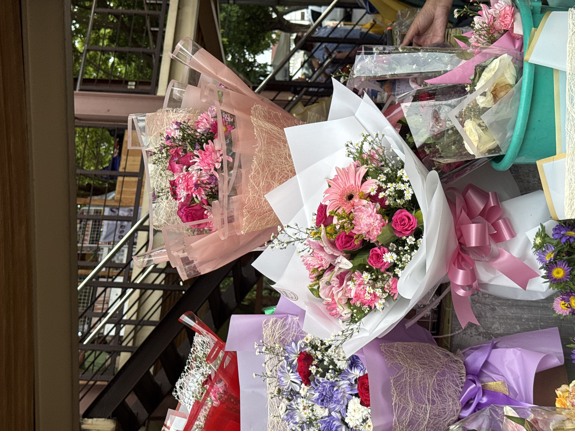 A close-up, medium shot captures a street vendor's stall in Davao City, Philippines, showcasing an assortment of floral bouquets. The bouquets are arranged prominently in the foreground, featuring a variety of colors including pink, red, purple, and white flowers. Some are wrapped in translucent plastic with decorative paper and ribbons, while others are presented in purple cellophane. In the middle ground, a set of metal stairs with a wooden railing is visible, suggesting an elevated walkway or building. The background is blurred, revealing glimpses of trees and foliage, indicating an outdoor setting within the city. A person's hand is seen reaching towards one of the bouquets in the right portion of the frame. The overall scene suggests a bustling urban environment where flowers are being sold.