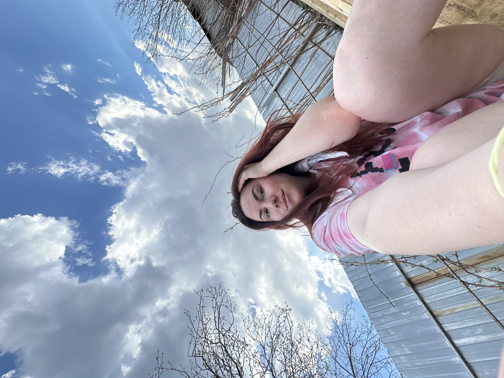 A low-angle shot shows a woman with long red hair, wearing a pink tie-dye shirt, posing against a backdrop of a blue sky with white clouds. The lower portion of her body is also visible. Behind her, there is a metal structure with bare branches. The location in Cheryomushki, Russia, cannot be determined from the image.