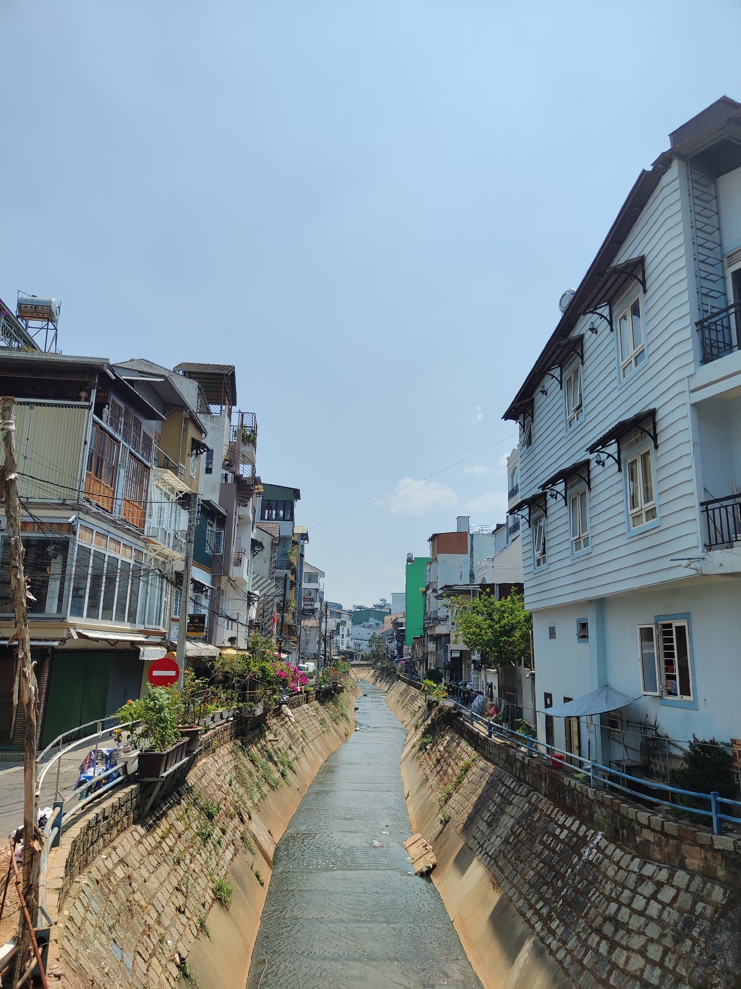 The image depicts a canal running through the city of Thành phố Đà Lạt, Vietnam. The canal is concrete-lined and filled with water. Buildings line both sides of the canal, varying in height and architectural style, ranging from older multi-story structures with balconies to newer, brightly colored buildings. A "do not enter" sign is visible on the left side of the canal, along with potted plants and vegetation along the canal's edge. Wires and utility poles are present on the left side, with a clear sky visible above. Some people are visible along the canal's edge.