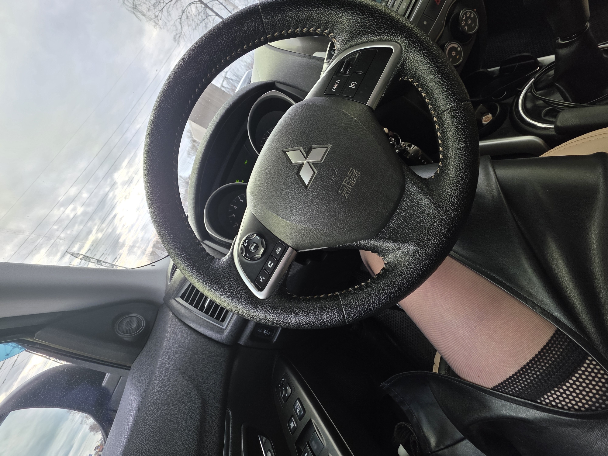 The image displays the interior of a Mitsubishi car, viewed from an upward angle, likely from the passenger side or a low perspective on the driver's side. The central focus is the black leather-wrapped steering wheel, featuring visible light-colored stitching. The Mitsubishi logo is prominently displayed in silver on the airbag cover, beneath which "SRS AIRBAG" is written. Controls for cruise control, including a "CANCEL" button, and other functions like audio/menu navigation are visible on the steering wheel.
A person's leg is partially visible beneath the steering wheel, indicating someone is seated in the driver's position. The leg is covered in light-colored hosiery or tights, topped with a distinct black patterned band, possibly fishnet, near the thigh. A black boot or shoe is visible on the foot, and a black, leather-like garment (possibly a skirt or dress) covers the upper leg.
Through the windshield, an overcast or cloudy sky is discernible, suggesting daytime. Bare tree branches and power lines are visible against the sky, along with a distant power pole or similar structure. Other parts of the car interior include air vents, a section of the dashboard, the instrument cluster, a speaker on the door panel, and window controls. The gear shift area, showing some cables, is also partially visible. The scene depicts someone sitting in a parked car or waiting inside it. This setting is located in Chaikovsky, Russia.